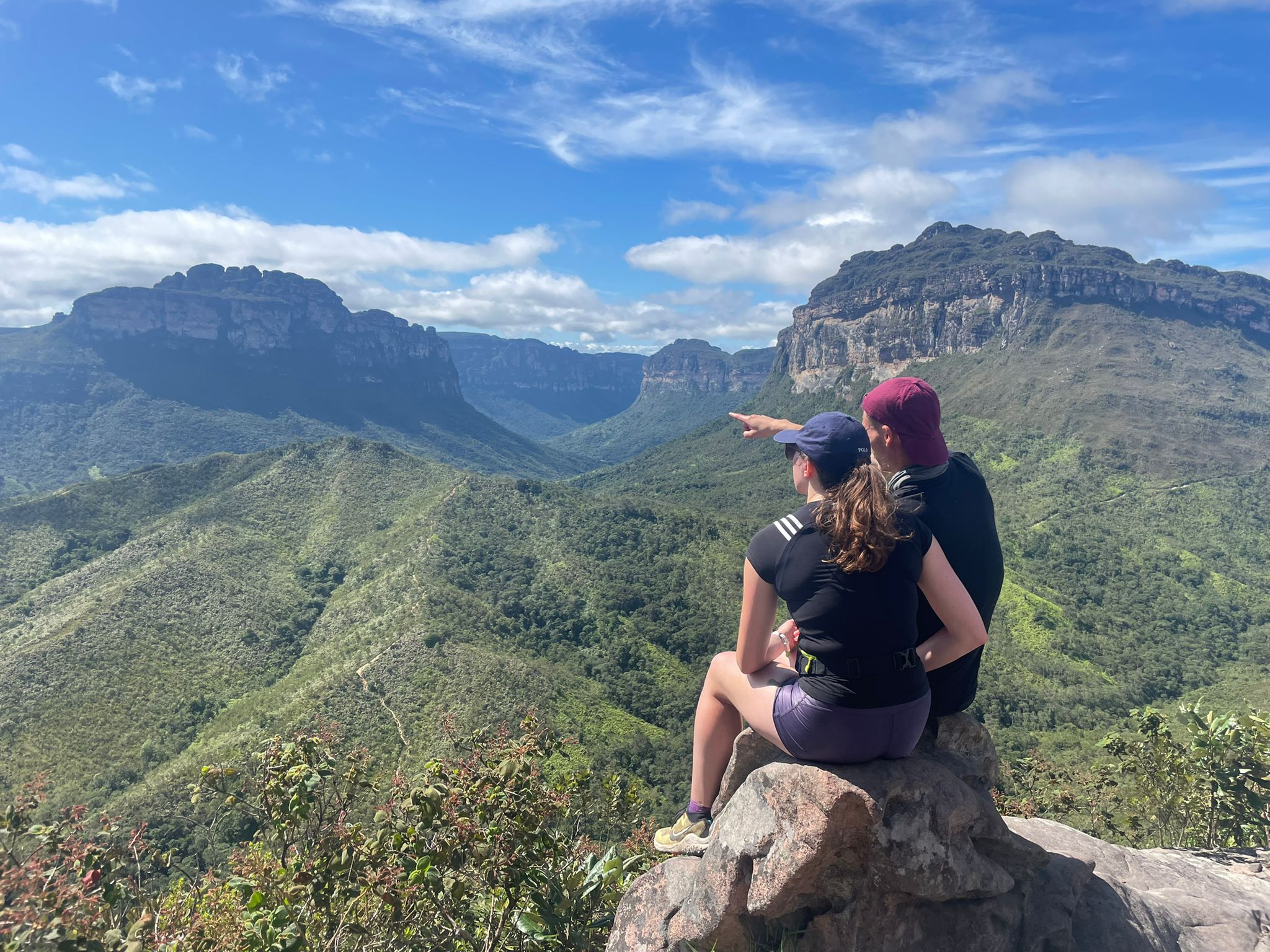 Two hikers sitting on a rock at Mirante do Pati, pointing towards the towering peaks of Morro do Castelo and Morro Branco. Two hikers sitting on a rock at Mirante do Pati, pointing towards the towering peaks of Morro do Castelo and Morro Branco.