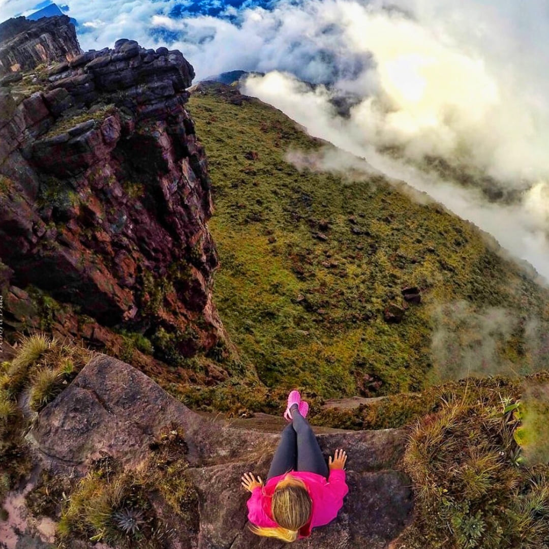 A hiker sits on the edge of a steep, rocky sandstone cliff on Mount Roraima, looking down at a vast valley filled with thick white clouds. A hiker sits on the edge of a steep, rocky sandstone cliff on Mount Roraima, looking down at a vast valley filled with thick white clouds.