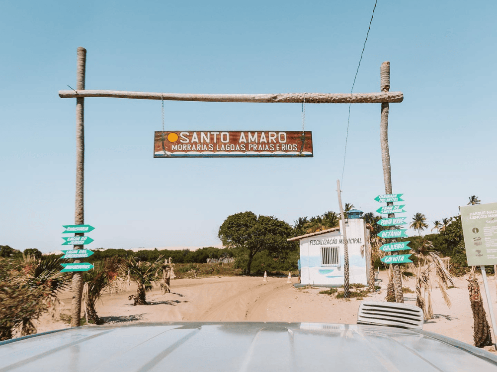 Rustic portico with a wooden sign displaying the text Santo Amaro Morvanas Lagoas Prajasthios over a sandy road. Rustic portico with a wooden sign displaying the text Santo Amaro Morvanas Lagoas Prajasthios over a sandy road.