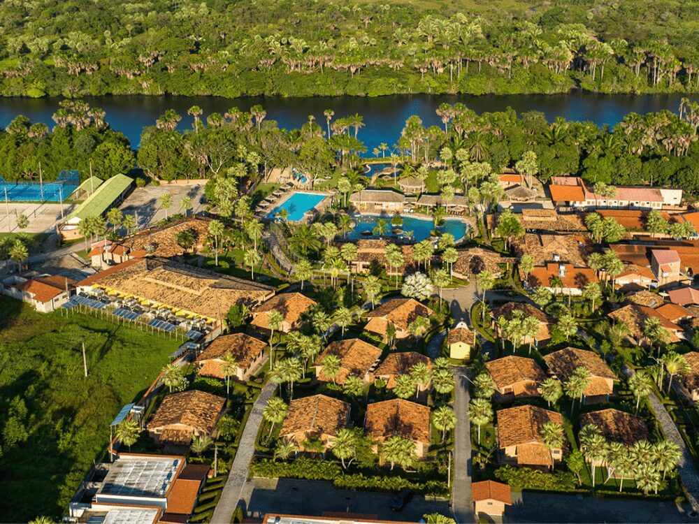 Aerial view of a lush resort complex featuring red-roofed buildings and blue swimming pools nestled beside a wide river.