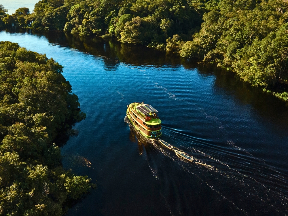 Aerial view of an Amazon river cruise boat navigating a dark river surrounded by dense green rainforest in Brazil. Aerial view of an Amazon river cruise boat navigating a dark river surrounded by dense green rainforest in Brazil.