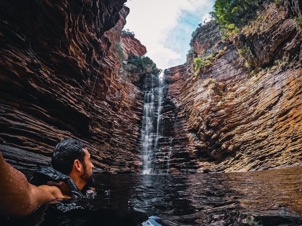 A man swims in a dark natural pool, looking up at a tall waterfall cascading down a canyon with layered rock formations. A man swims in a dark natural pool, looking up at a tall waterfall cascading down a canyon with layered rock formations.
