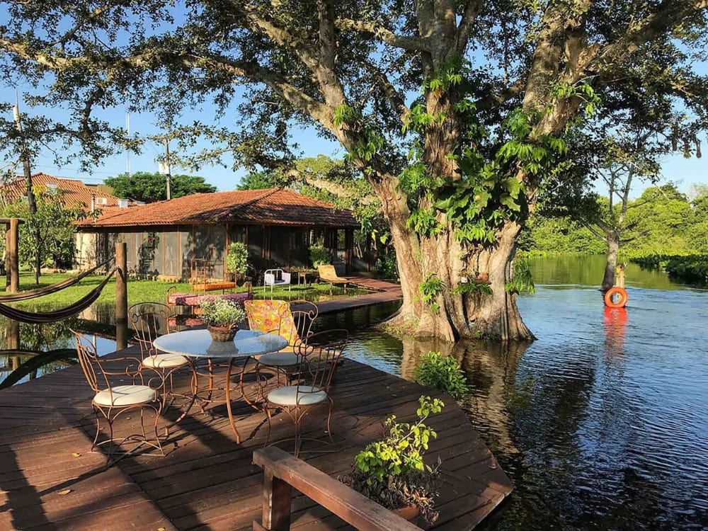 A wooden deck with a table and chairs sits beside a tranquil lake and a massive tree at RefĂșgio da Ilha ecolodge. A wooden deck with a table and chairs sits beside a tranquil lake and a massive tree at RefĂșgio da Ilha ecolodge.