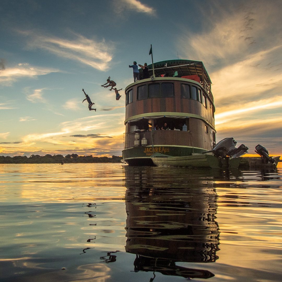 Tourists jumping into the river from the top deck of the Jacaré-Açu wooden boat at sunset during an authentic Amazon river cruise experience.