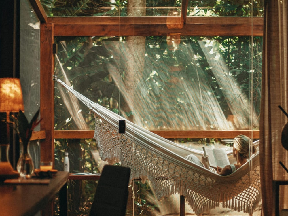  A person relaxing in a hammock inside a sophisticated room with large glass windows facing the jungle.