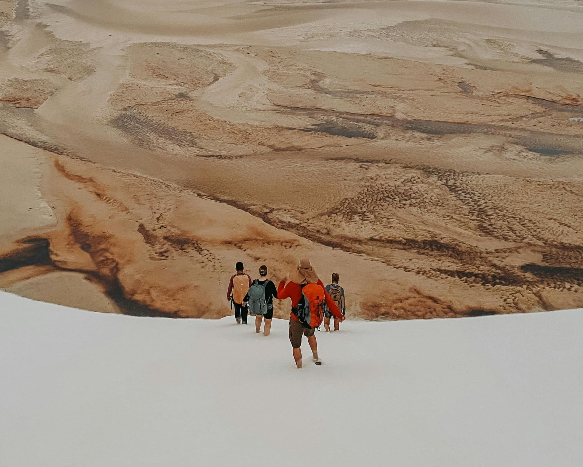  A group of tourists walks across a dry, brown lagoon bed surrounded by large white sand dunes during the dry season.