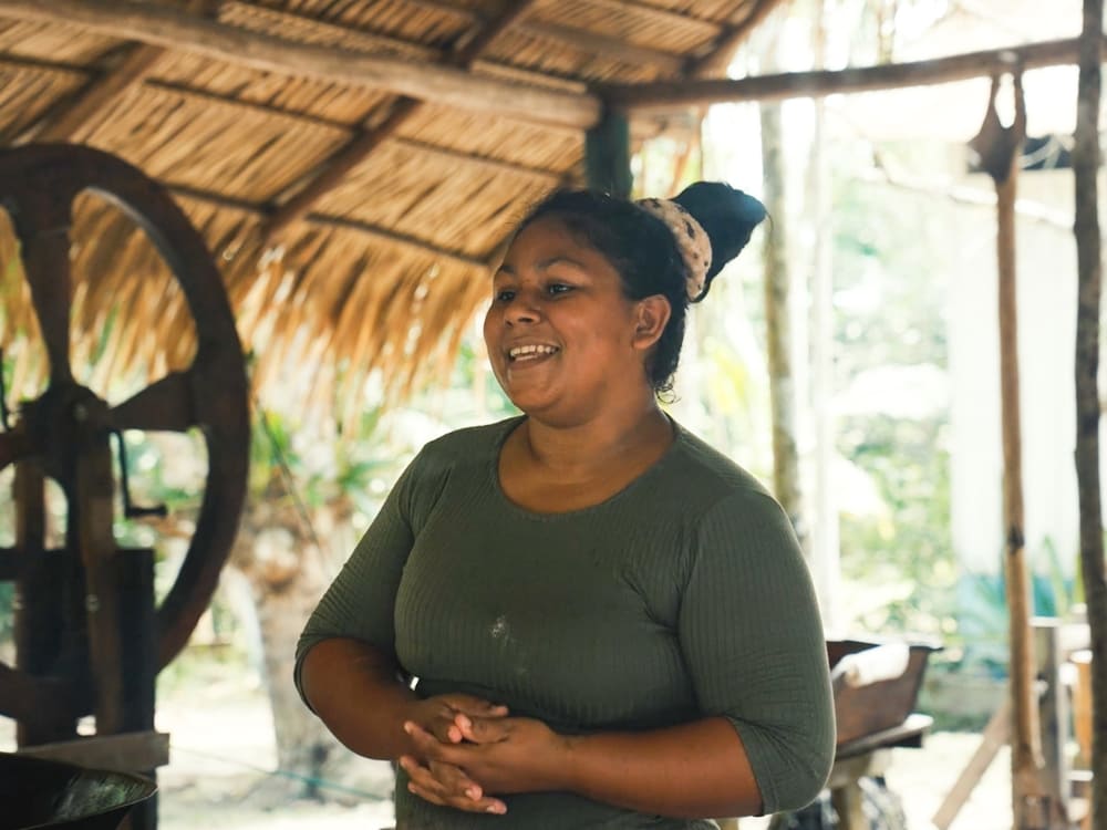 Smiling woman from an Amazon riverside community standing under a traditional thatched roof.