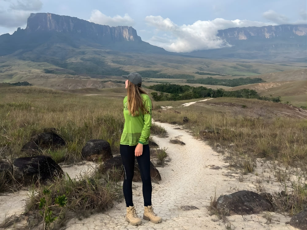 A female ecologist stands on a sandy path looking toward the table mountain of Mount Roraima in the distance.