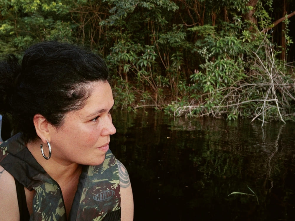A dark-haired woman wearing hoop earrings and a camouflage life vest sits in a small boat, looking thoughtfully at the lush riverbank vegetation