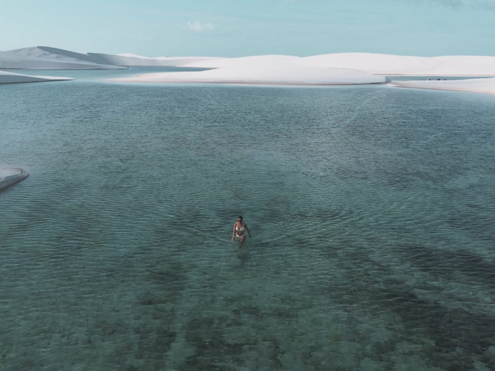 Woman walking in the shallow, crystal-clear waters of a lagoon surrounded by the large white sand dunes of Lençóis Maranhenses.