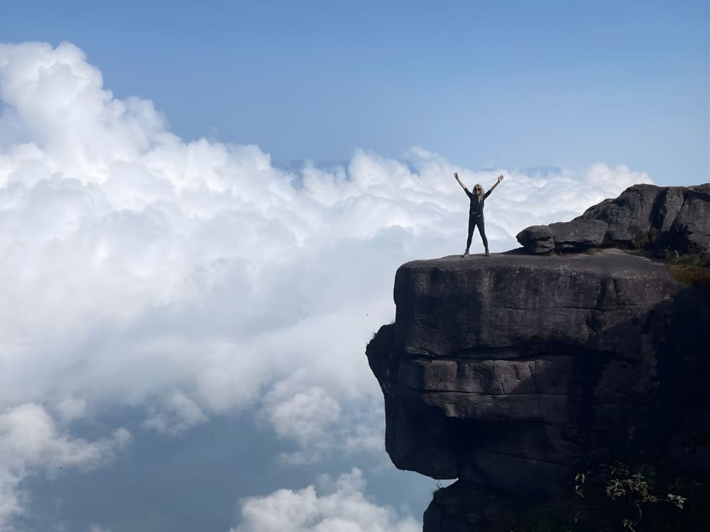 Woman with raised arms on top of a large rocky cliff at Mount Roraima, celebrating surrounded by dense clouds.
