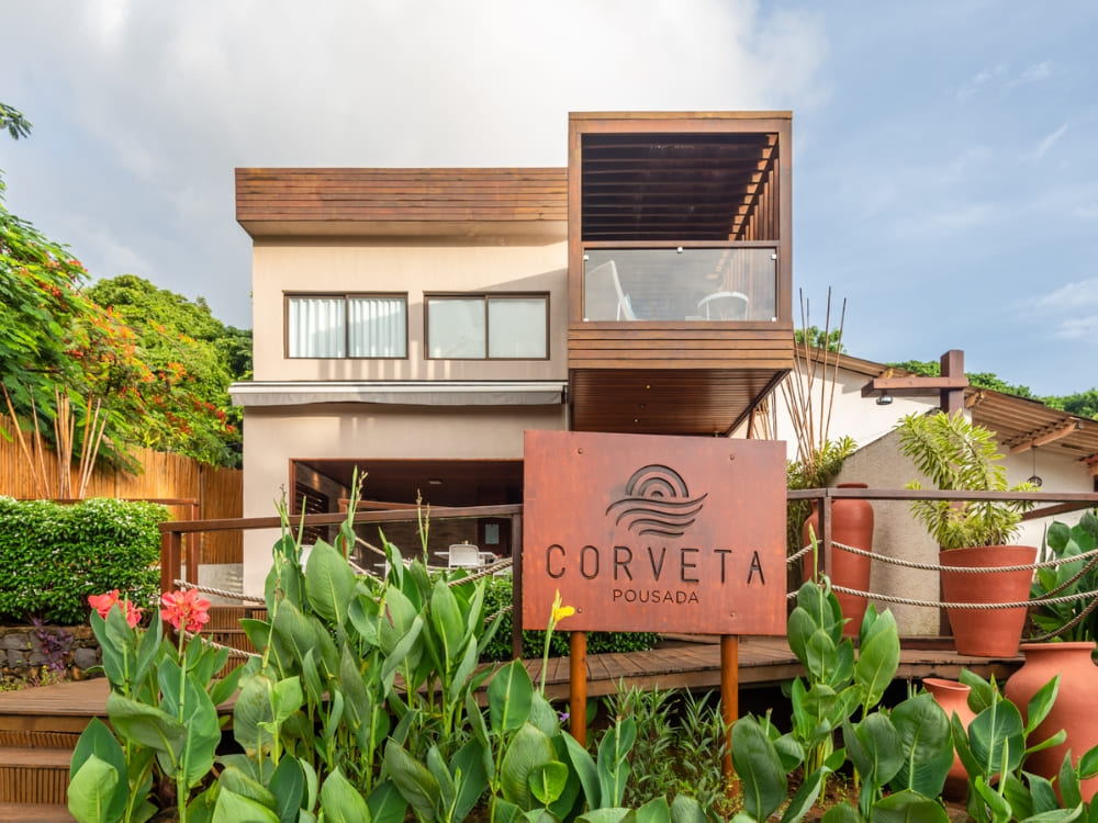 Modern exterior of Pousada Corveta with a metal sign, wooden walkways, and tropical plants, highlighting where to stay in Fernando de Noronha. Modern exterior of Pousada Corveta with a metal sign, wooden walkways, and tropical plants, highlighting where to stay in Fernando de Noronha.