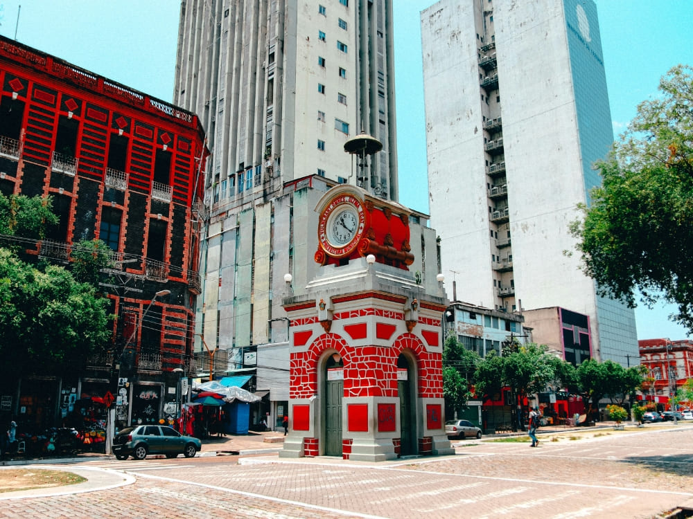 Historic red and stone clock tower standing in a city square against a backdrop of modern tall buildings in Manaus. Historic red and stone clock tower standing in a city square against a backdrop of modern tall buildings in Manaus.
