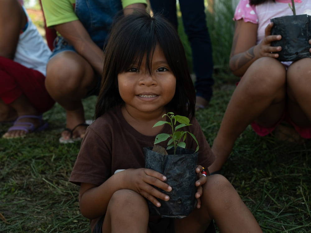 A smiling child from an Amazonian riverside community sitting on the grass and holding a small potted plant. A smiling child from an Amazonian riverside community sitting on the grass and holding a small potted plant.
