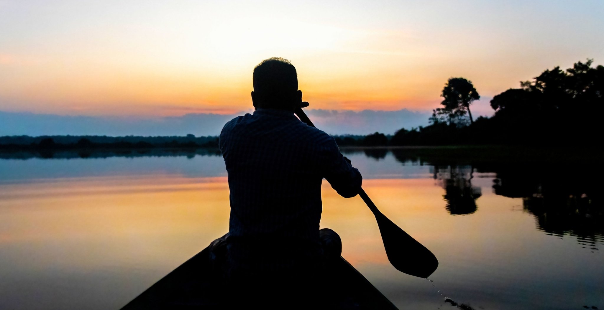 Silhouette eines Paddlers in einem Kanu auf einem spiegelglatten Fluss bei Sonnenuntergang, mit warmem orange-rosa Himmel, der sich im Wasser spiegelt, und dunkler Waldsilhouette am Amazon-Boutique-Lodge-Wasserweg