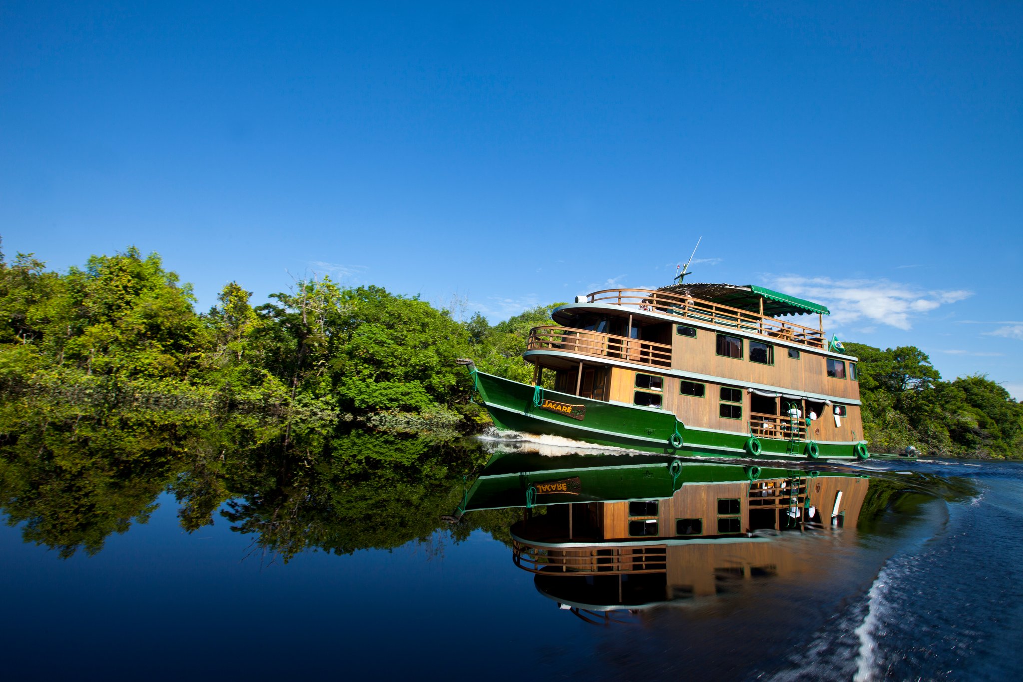 The Jacaré-Açu wooden Amazon cruise trip vessel in Brazil glides along a dark-water river channel flanked by dense rainforest, its green-and-wood hull perfectly mirrored in the glassy surface