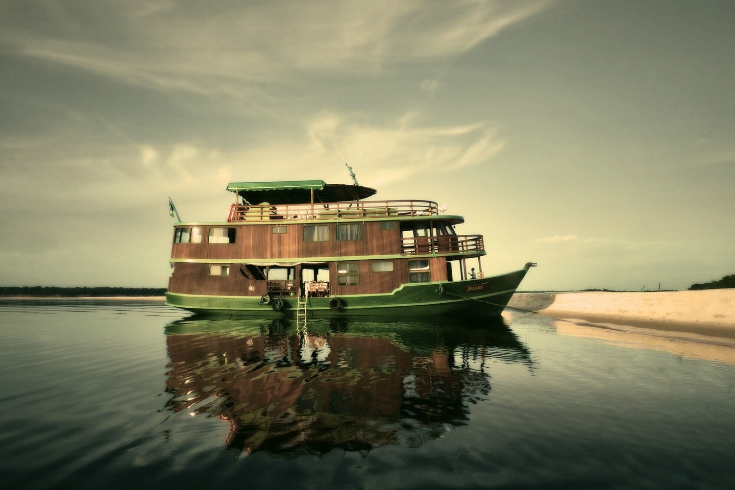 The Jacaré-Açu riverboat, a two-deck wooden vessel with green trim, moored on calm dark waters beside a white sandbank on an Amazon Cruise Trip in Brazil