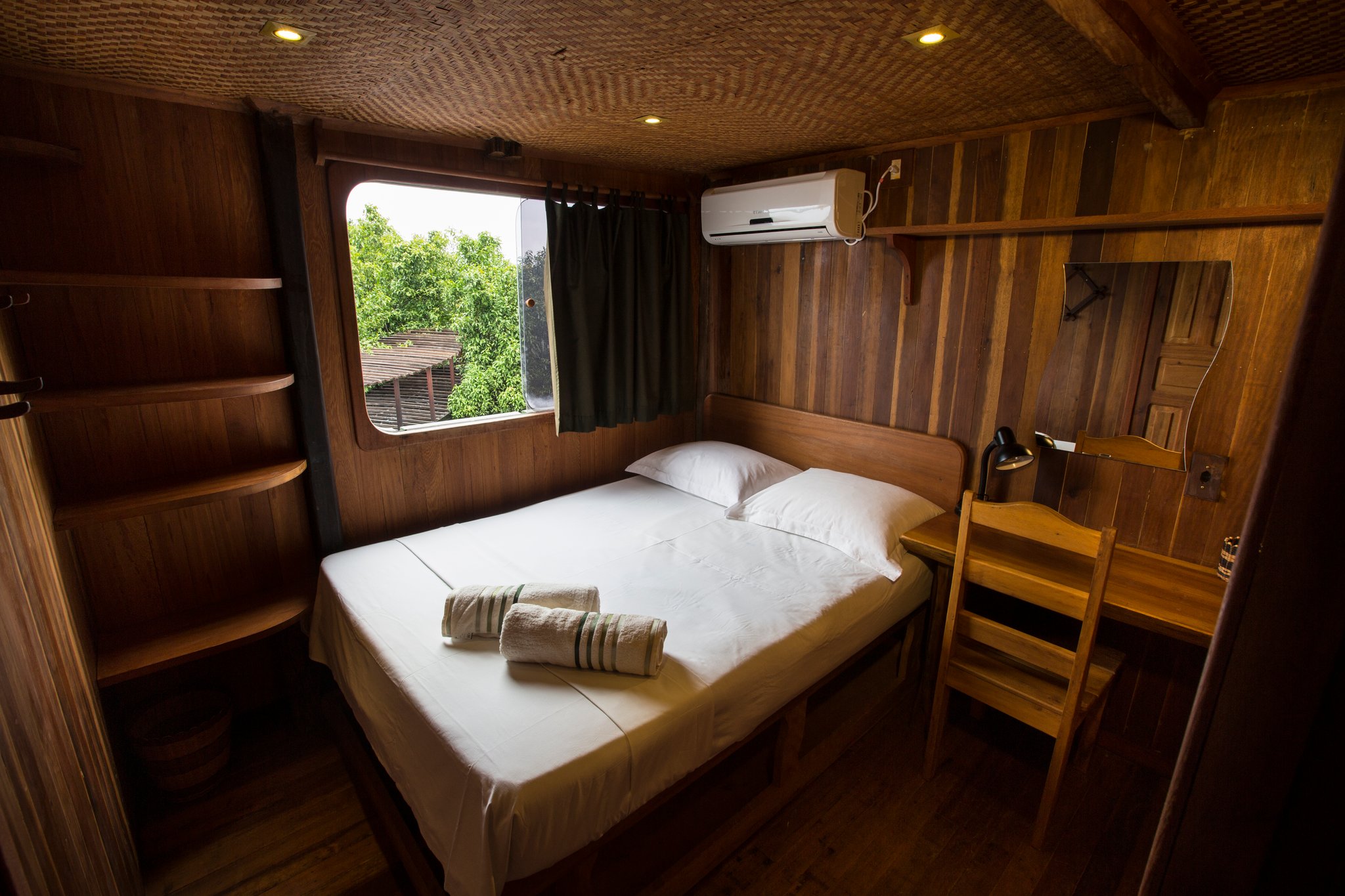 Small space, but the rainforest right outside the porthole makes up for it. Snug wood-panelled cabin on an Amazon jungle cruise Brazil vessel, featuring a double bed with white linens, rolled towels, built-in shelves, a desk, air conditioning, and a porthole-style window overlooking greenery