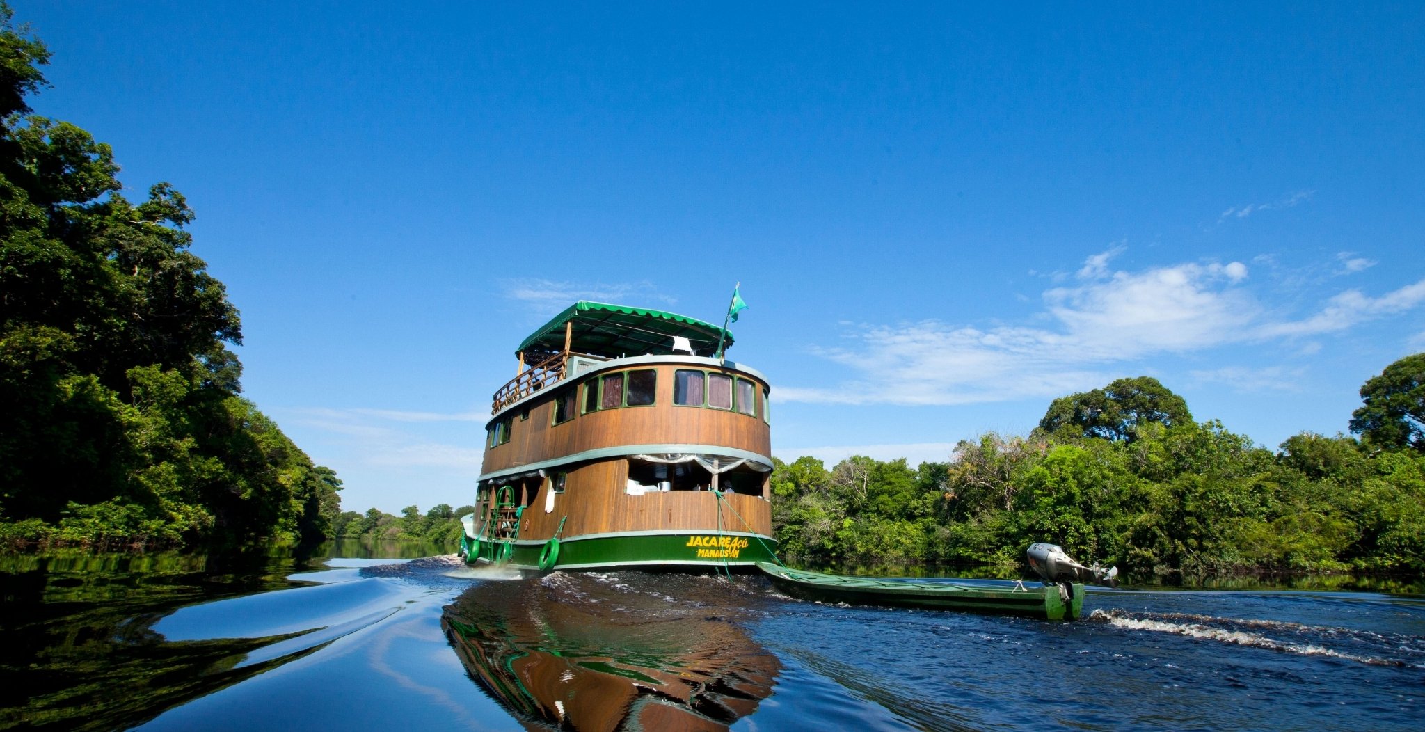 The Jacaré-Açu wooden riverboat cruising a dark-water Amazon jungle cruise Brazil channel flanked by dense rainforest under a vivid blue sky