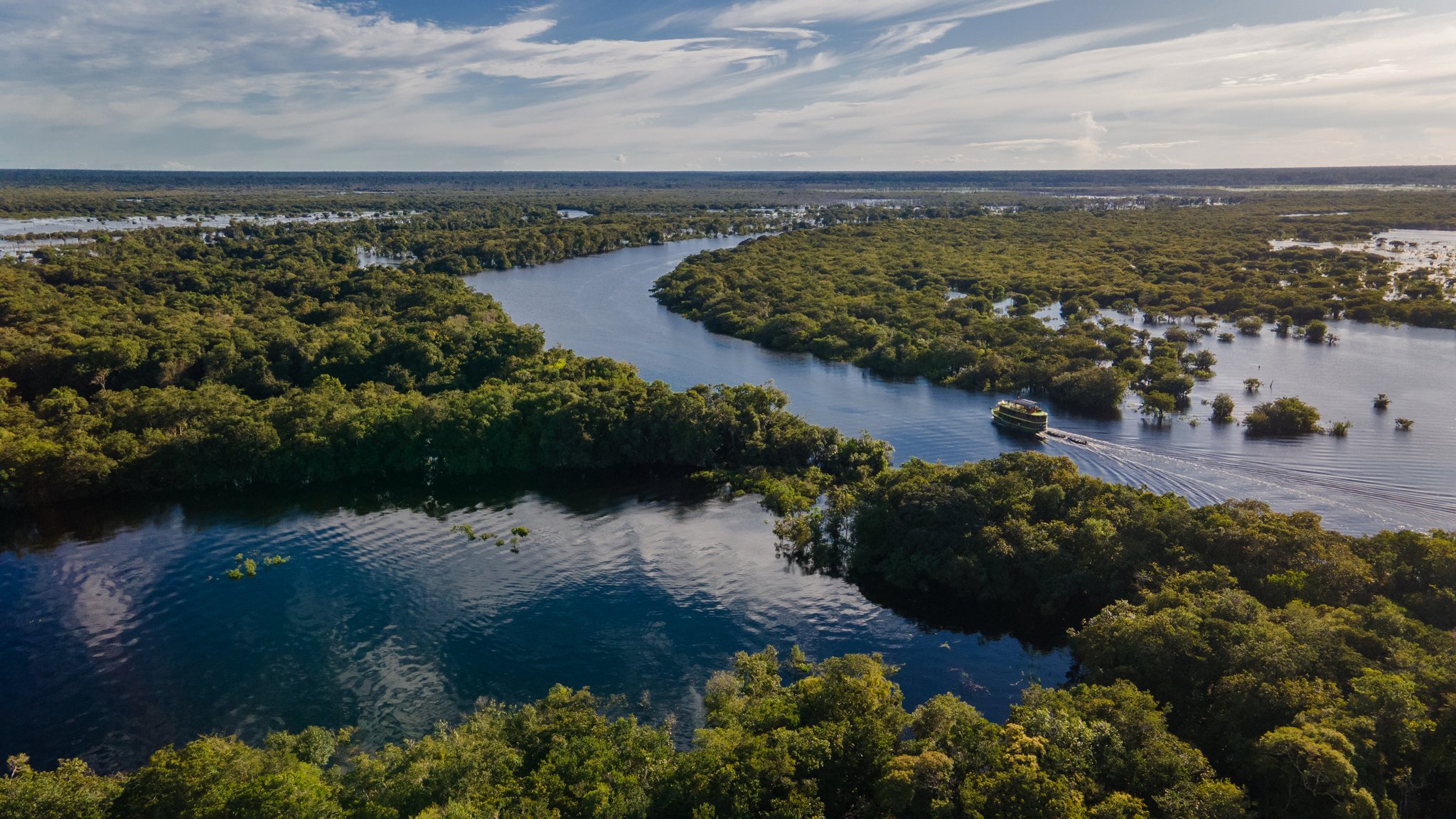 Aerial view of a riverboat navigating a winding river channel through flooded igapó forest on an Amazon river cruise in Brazil, with tree canopies stretching to the horizon under a partly cloudy sky
