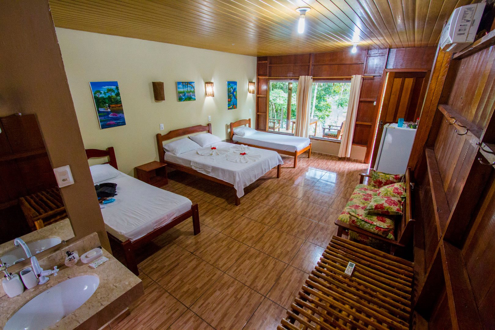 Netjes en rustig, met het oerwoud dat zachtjes op de ruit klopt. Ruime driepersoonskamar in het Amazon Turtle Lodge met drie houten bedden in wit linnengoed, tropische schilderijen aan crèmewitte muren en een raam met uitzicht op het regenwoudkronendak