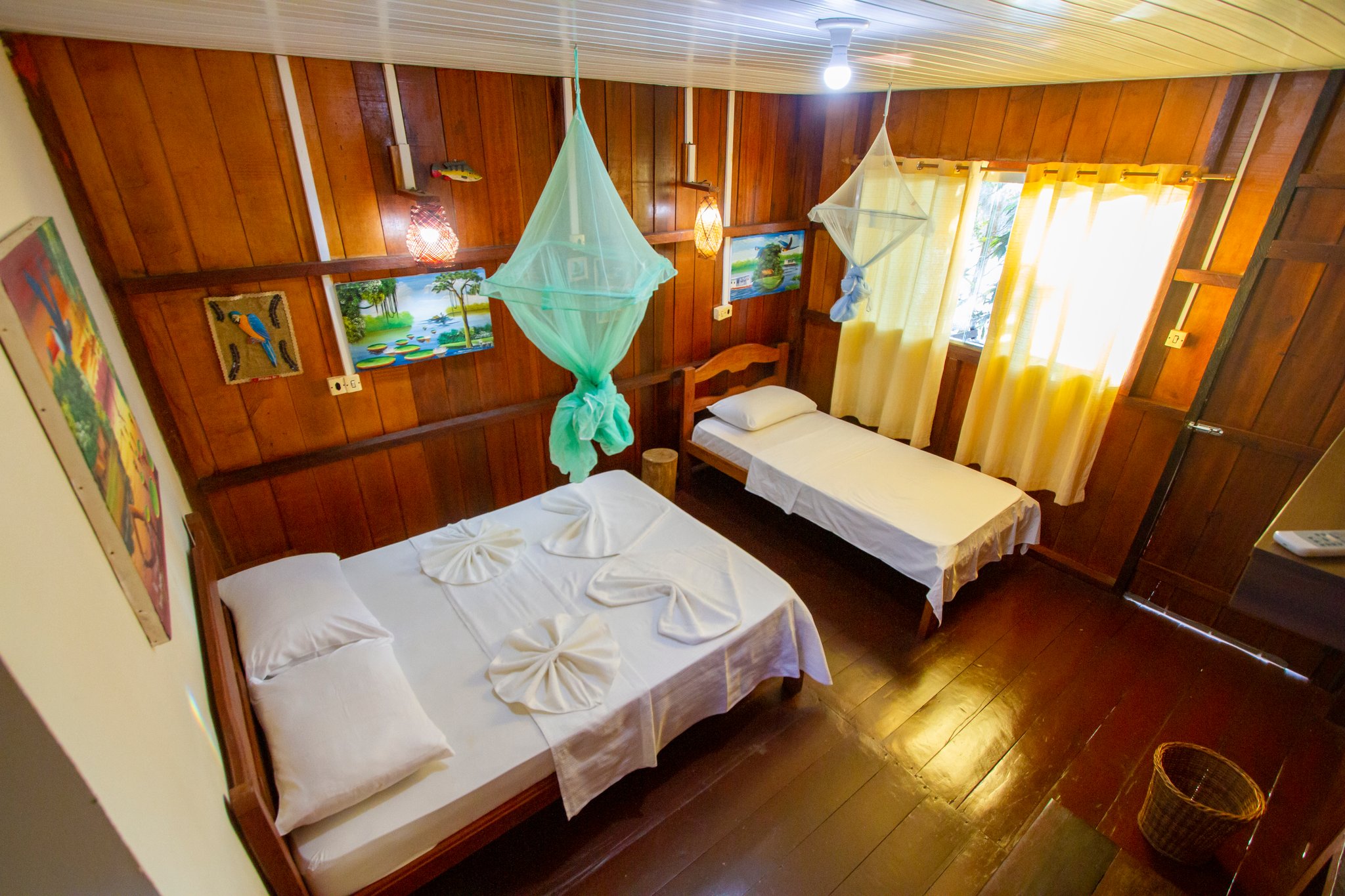 Klein maar fijn, na een dag diep in het regenwoud. Houten tweepersoonskamer in Amazon Turtle Lodge met een tweepersoonsbed en een eenpersoonsbed, groene en witte klamboes, gevouwen handdoekenkunst op witte beddengoed en kleurrijke regenwoudschilderijen aan de wanden