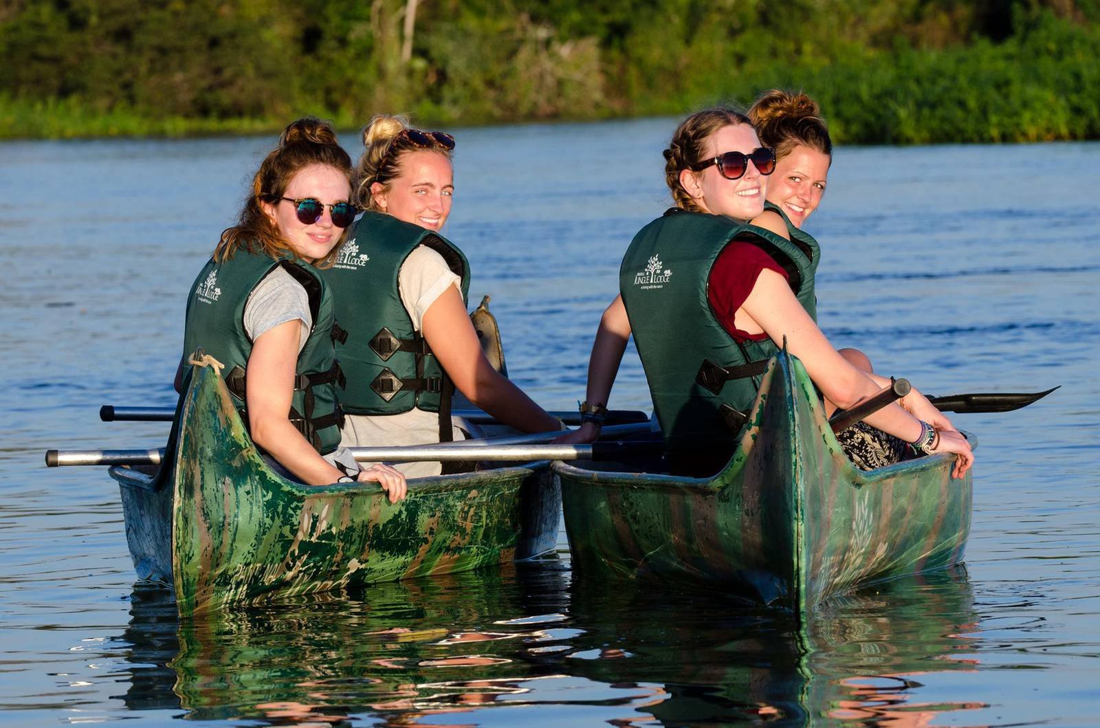 Vier lachende vrouwen in groene zwemvesten peddelen in een kano op een rustige rivier tijdens een goedkope Pantanal-tour, met dicht groen bos op de achtergrond