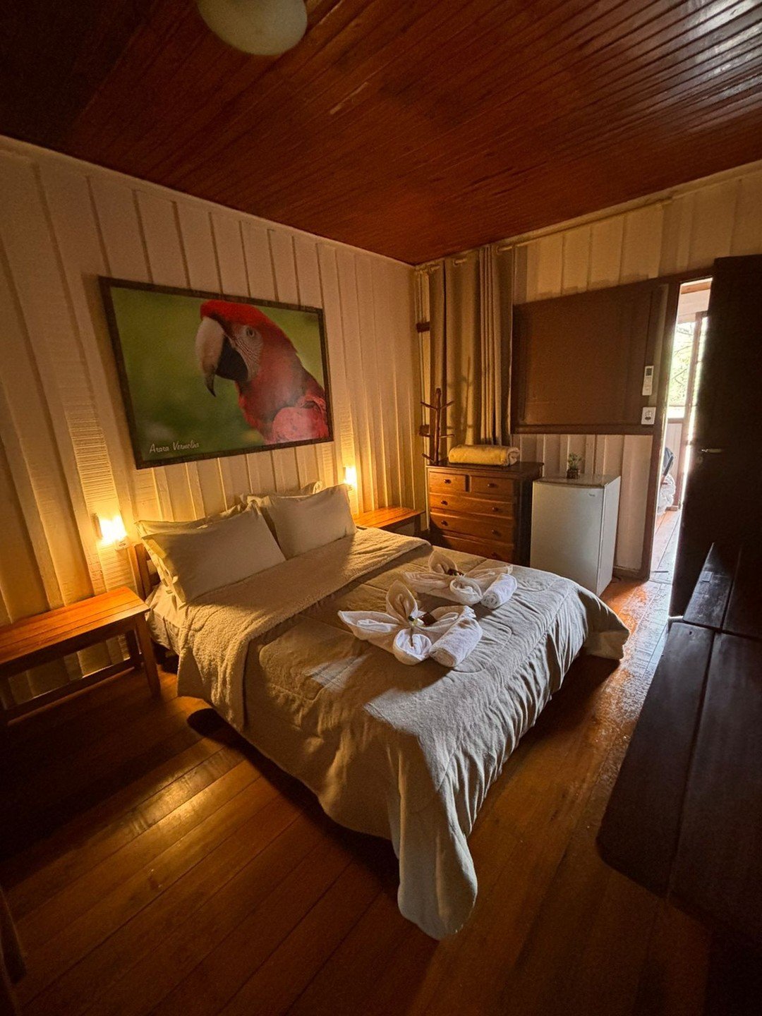 Gezellige tweepersoonskamer in een betaalbare Pantanal-lodge met donker houten plafond, zacht sfeerverlichting, decoratief gevouwen handdoeken op het bed, een houten ladekast, minikoelkast en een ingelijst portret van een rode ara aan de witte houten wand