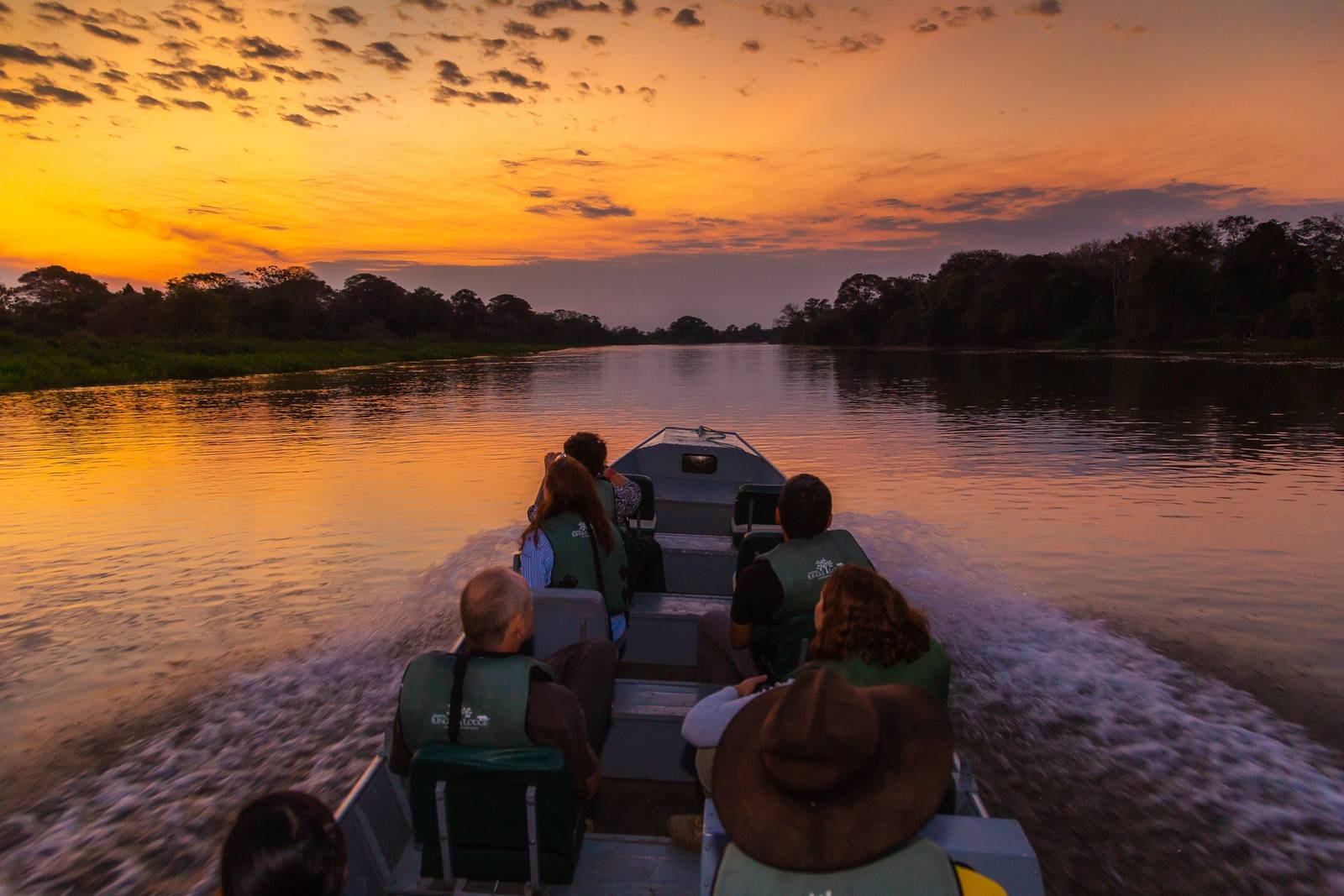 Reizigers in zwemvesten op een motorboot die bij een feloranje zonsondergang over een rustig rivierkanaal in het Pantanal vaart, met bossen als silhouet aan beide oevers
