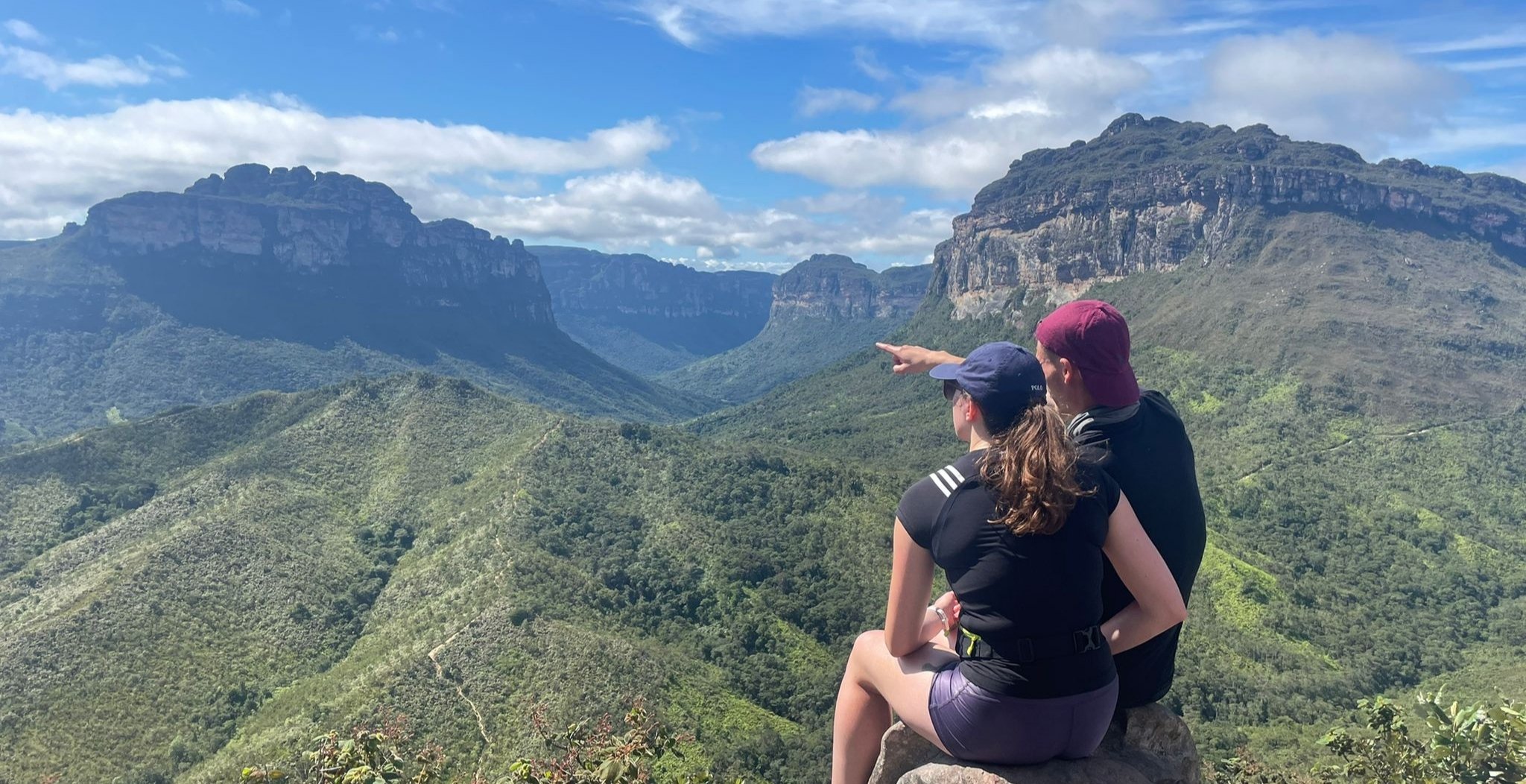 Zwei Wanderer sitzen auf einem Felsvorsprung beim Chapada-Diamantina-Trek und zeigen auf das weite, bewaldete Tal des Vale do Pati, flankiert von mächtigen Sandsteinplateaus unter blauem Himmel