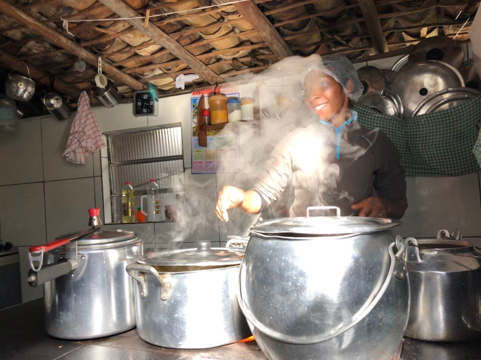 A smiling cook wearing a hairnet tends large steaming aluminium pots in a rustic kitchen with a wooden-beam ceiling, an authentic glimpse into community based tourism hospitality in Brazil
