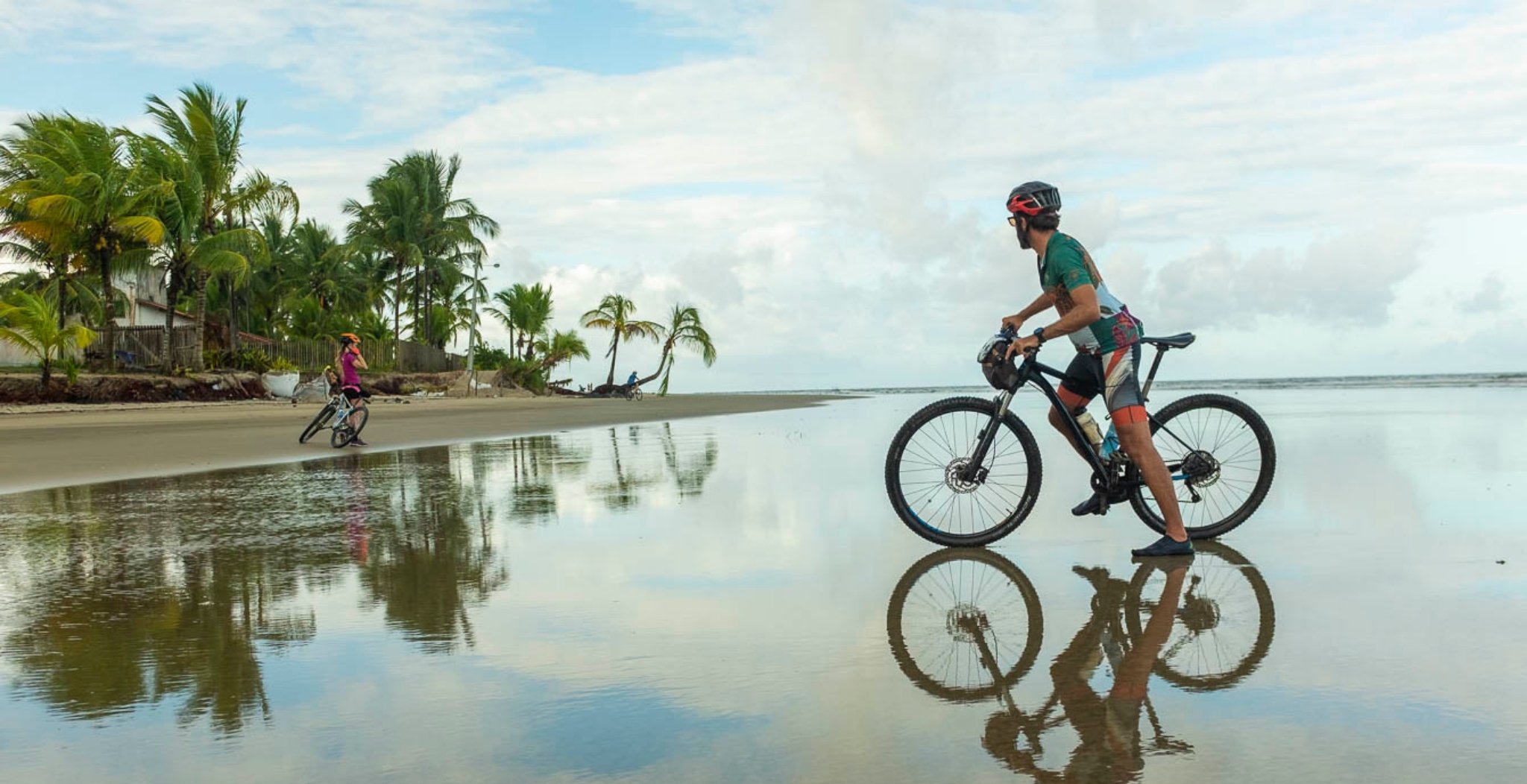 Three cyclists riding mountain bikes along a wide, mirror-flat low-tide beach lined with coconut palms during a cycling Bahia tour, their reflections perfectly doubled in the wet sand