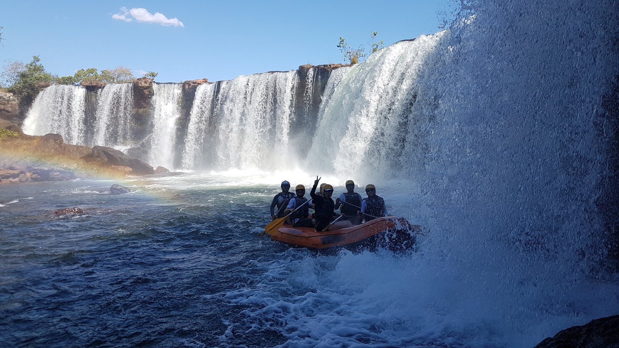 A group of helmeted rafters in an orange inflatable raft navigating the churning whitewater at the base of a wide horseshoe waterfall during a Jalapão trekking tour, with a rainbow forming in the mist to the left