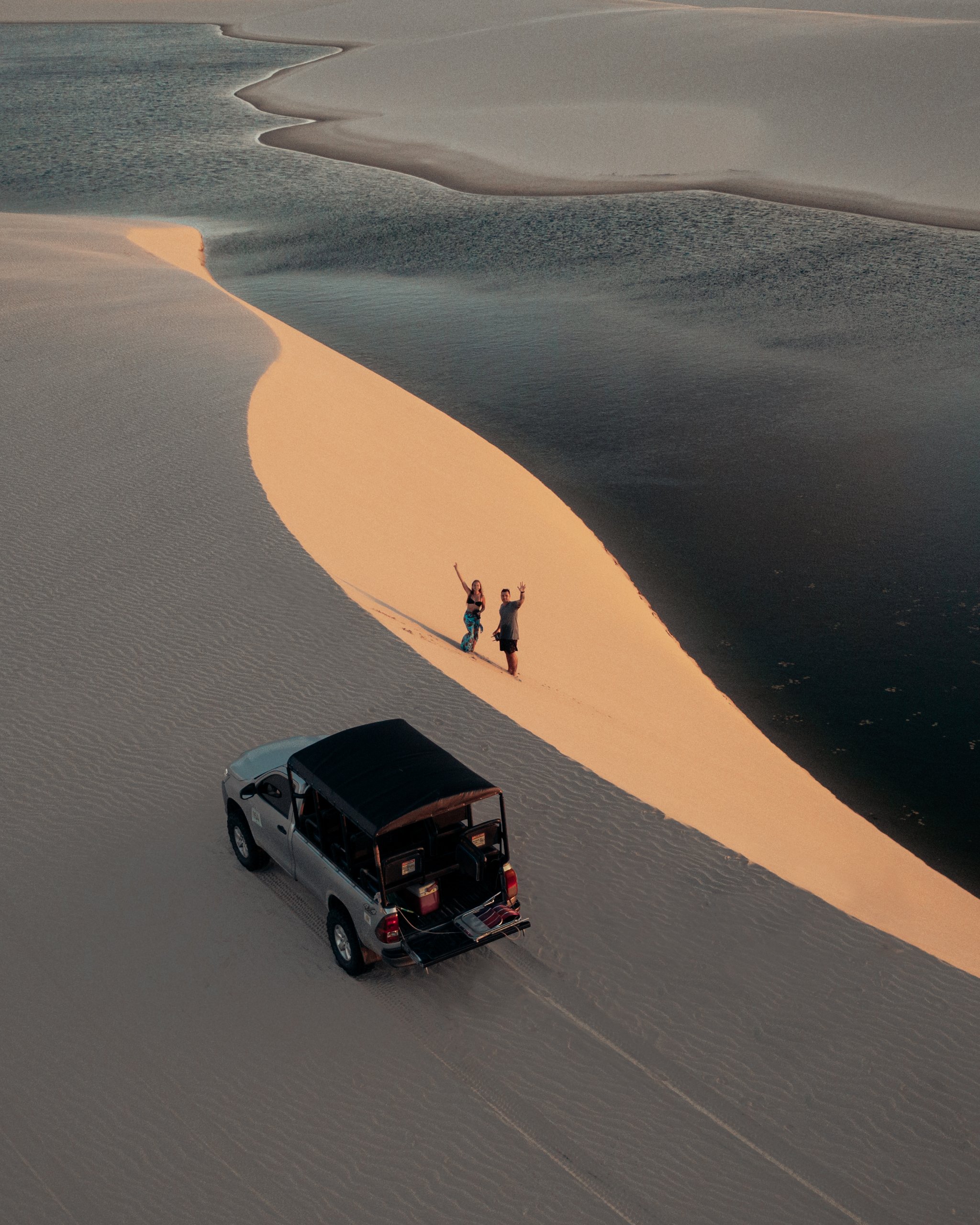 Aerial view of two travelers waving from a golden sand dune ridge beside a dark reflective lagoon during sunset, with a 4x4 vehicle parked below — a scene from a Lençóis Maranhenses full itinerary