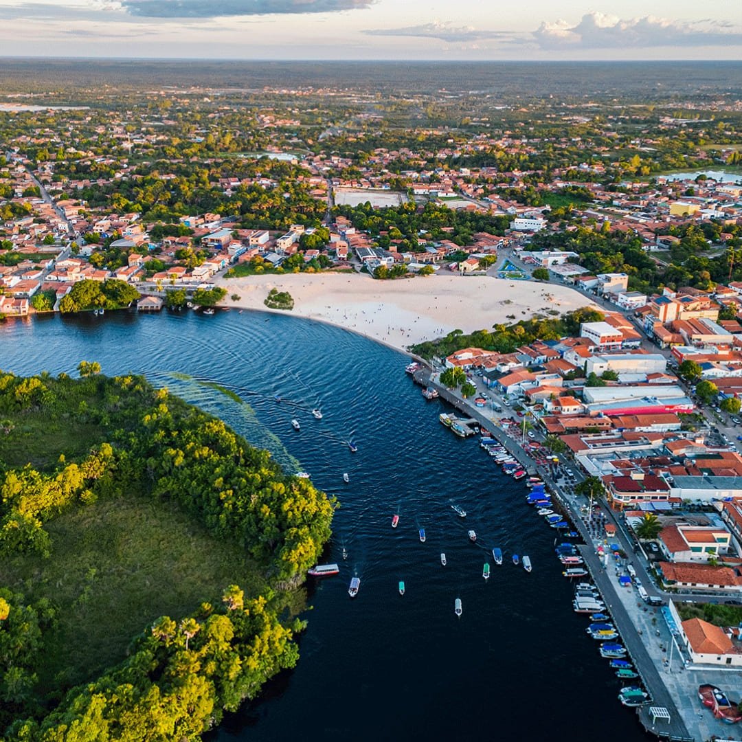 Aerial view of Barreirinhas showing the Preguiças River curving past a white sand beach, colorful boats moored along the waterfront, and the town spreading into green vegetation on the Lençóis Maranhenses Full Itinerary