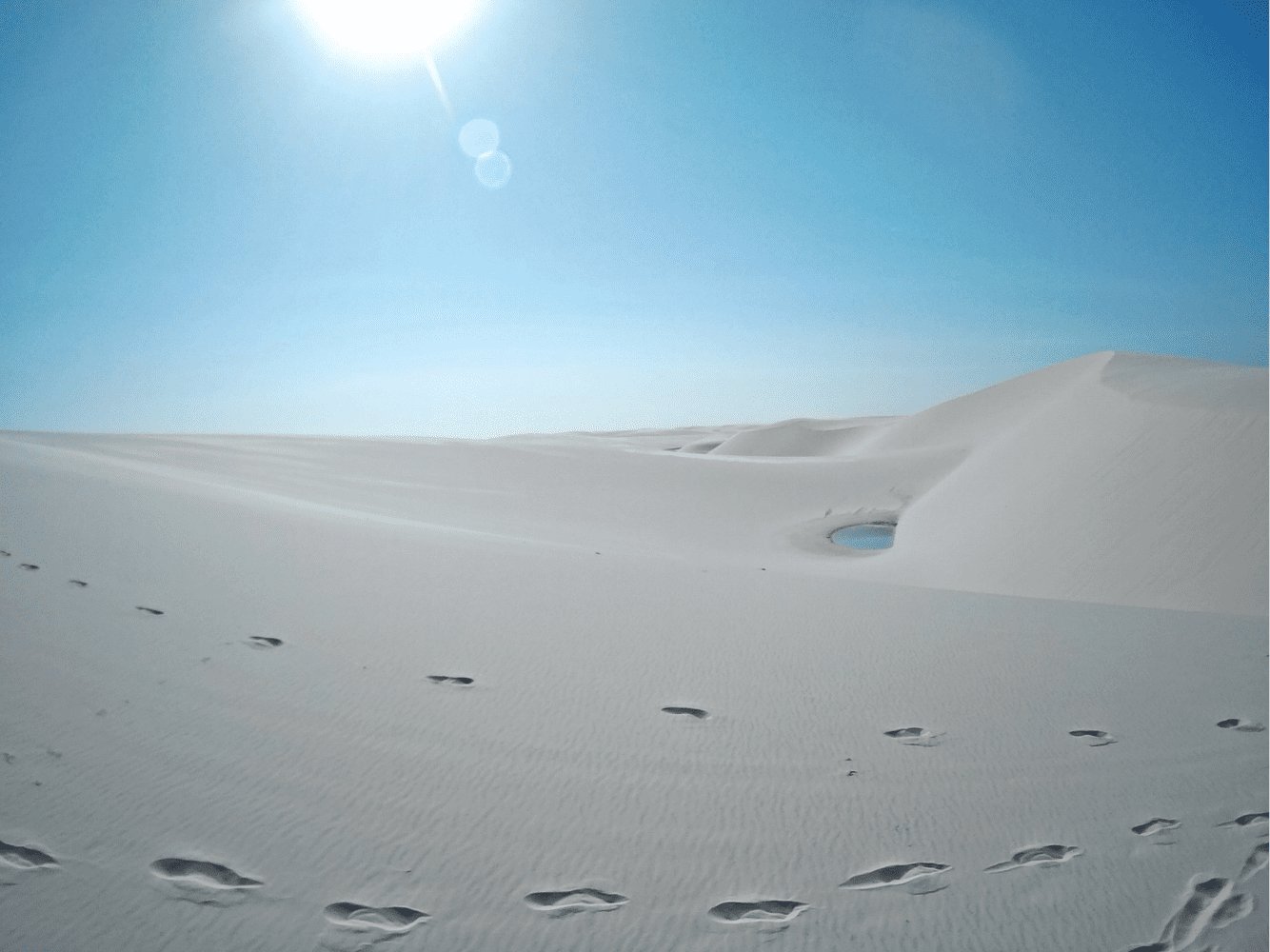 Bare footprints crossing vast white sand dunes toward a small blue lagoon nestled between ridges under a blazing sun — a scene from the Lençóis Maranhenses Full Itinerary