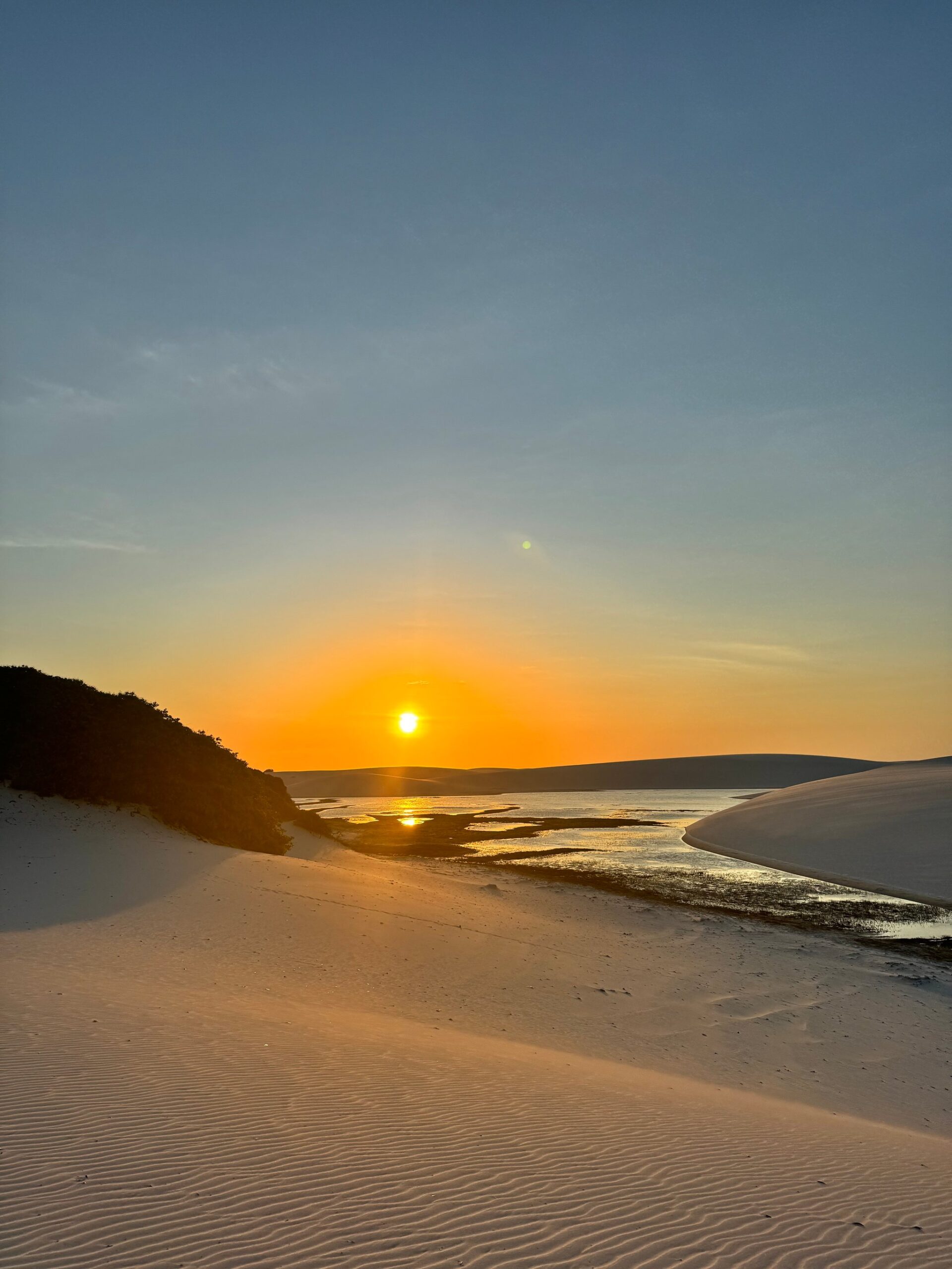 Golden sunset over a tidal channel winding between white sand dunes in Lençóis Maranhenses, reflecting orange light on shallow water