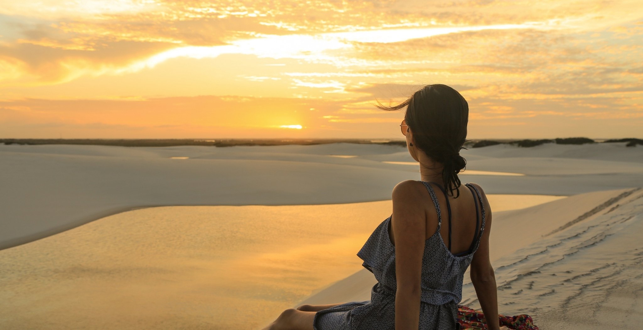 A woman sits alone on a white sand dune watching a vivid golden sunset over the Lençóis Maranhenses full itinerary landscape of dunes and shallow lagoons