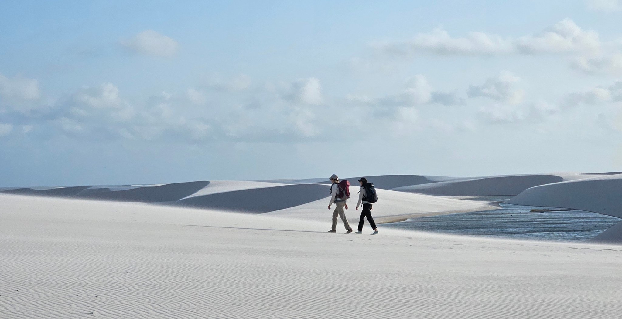 Two hikers with backpacks on a Lençóis Maranhenses hike, walking across vast white sand dunes beside a shallow lagoon under a pale blue sky