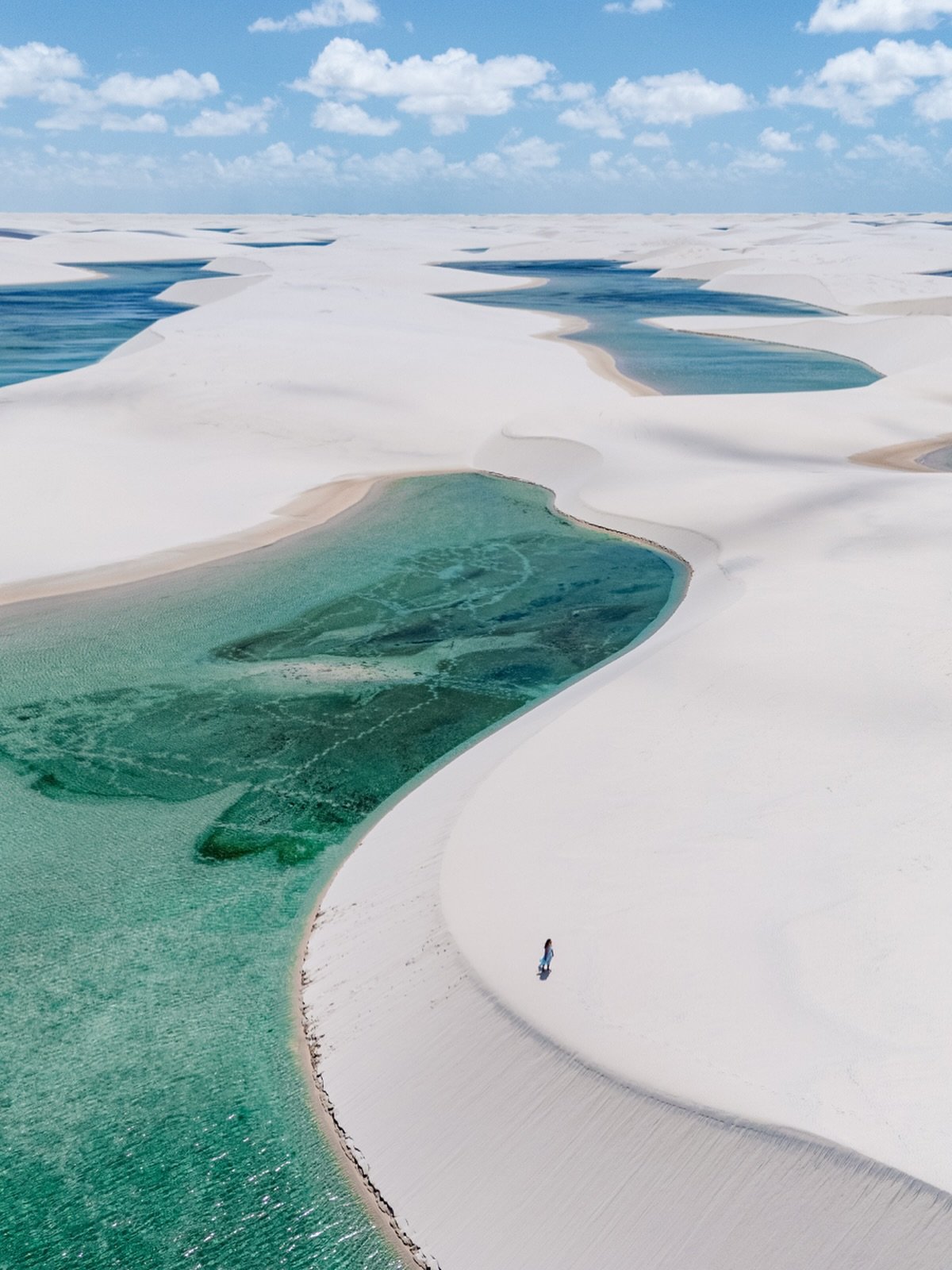 Luftaufnahme der Lençóis Maranhenses bei Santo Amaro mit weitläufigen weißen Sanddünen, türkis- und smaragdfarbenen Regenwasserlagune bis zum Horizont und einer einzelnen Person am Ufer unter blauem Himmel