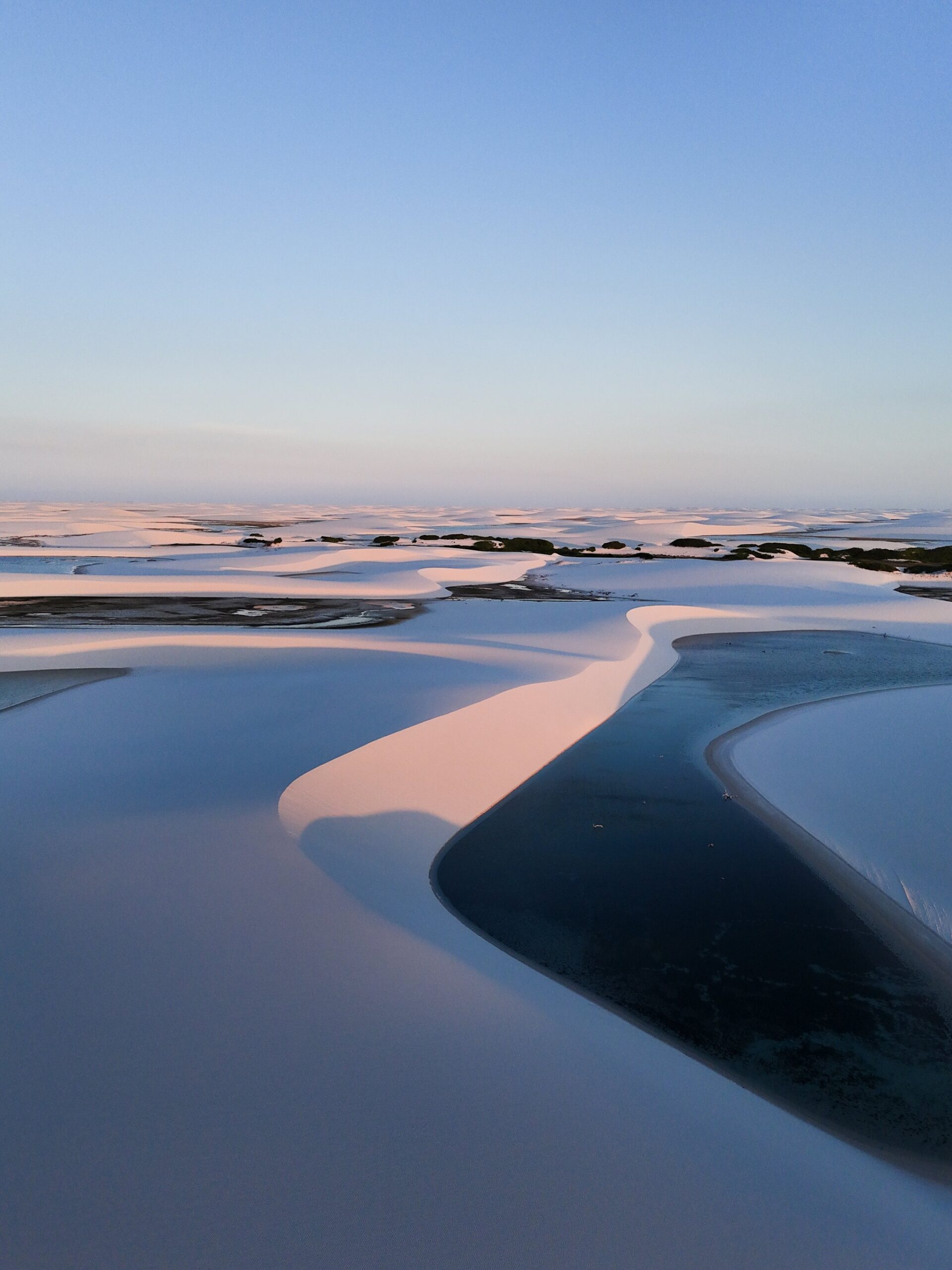 Luftaufnahme der weißen Sanddünen von Lençóis Maranhenses bei Dämmerung, die im rosafarbenen Abendlicht leuchten, während sich dunkle Süßwasserlagune zwischen ihnen schlängeln
