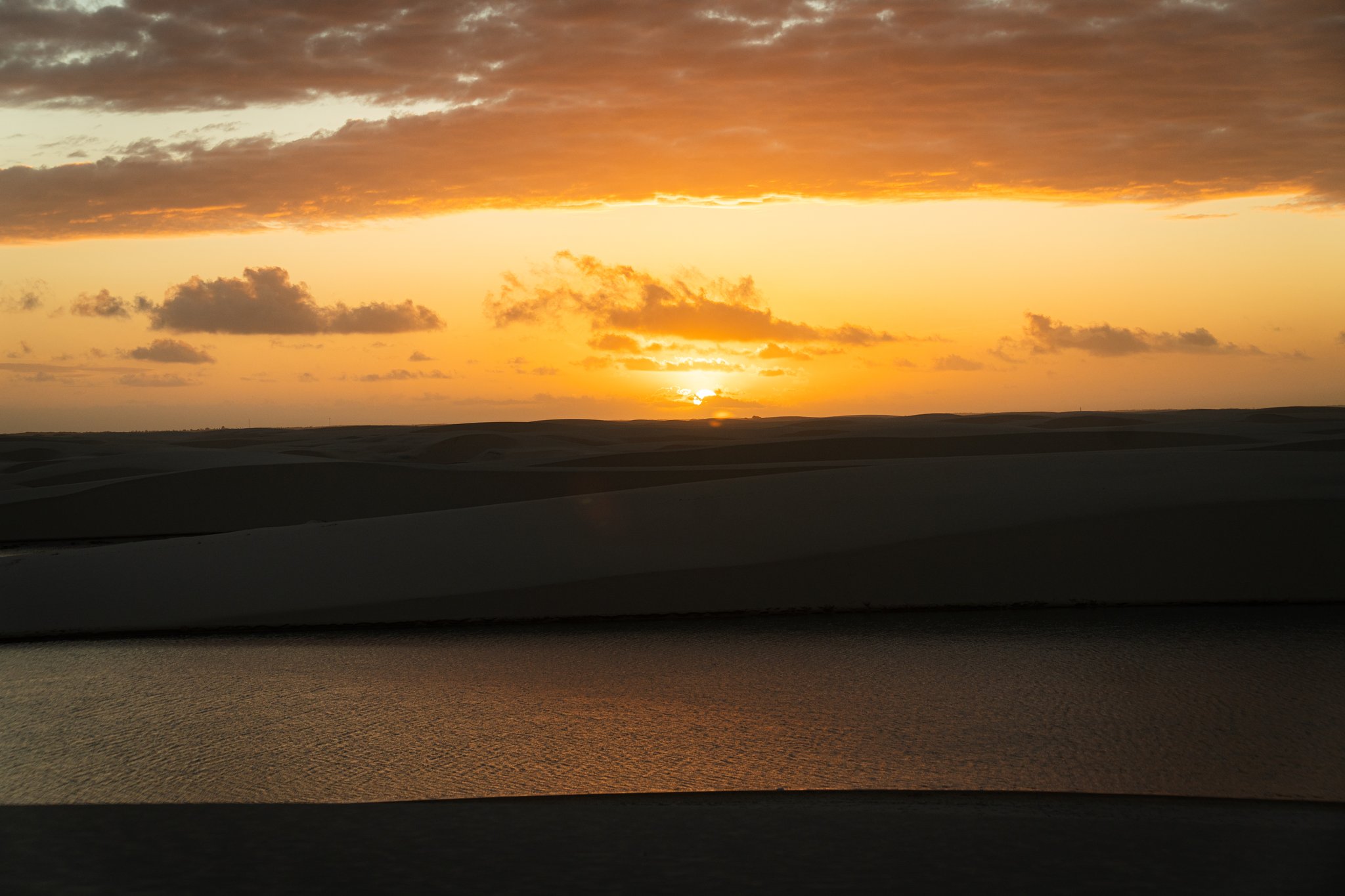 Golden sunset over silhouetted sand dunes and a calm reflective lagoon in Lençóis Maranhenses Santo Amaro, with orange clouds stretching across the sky