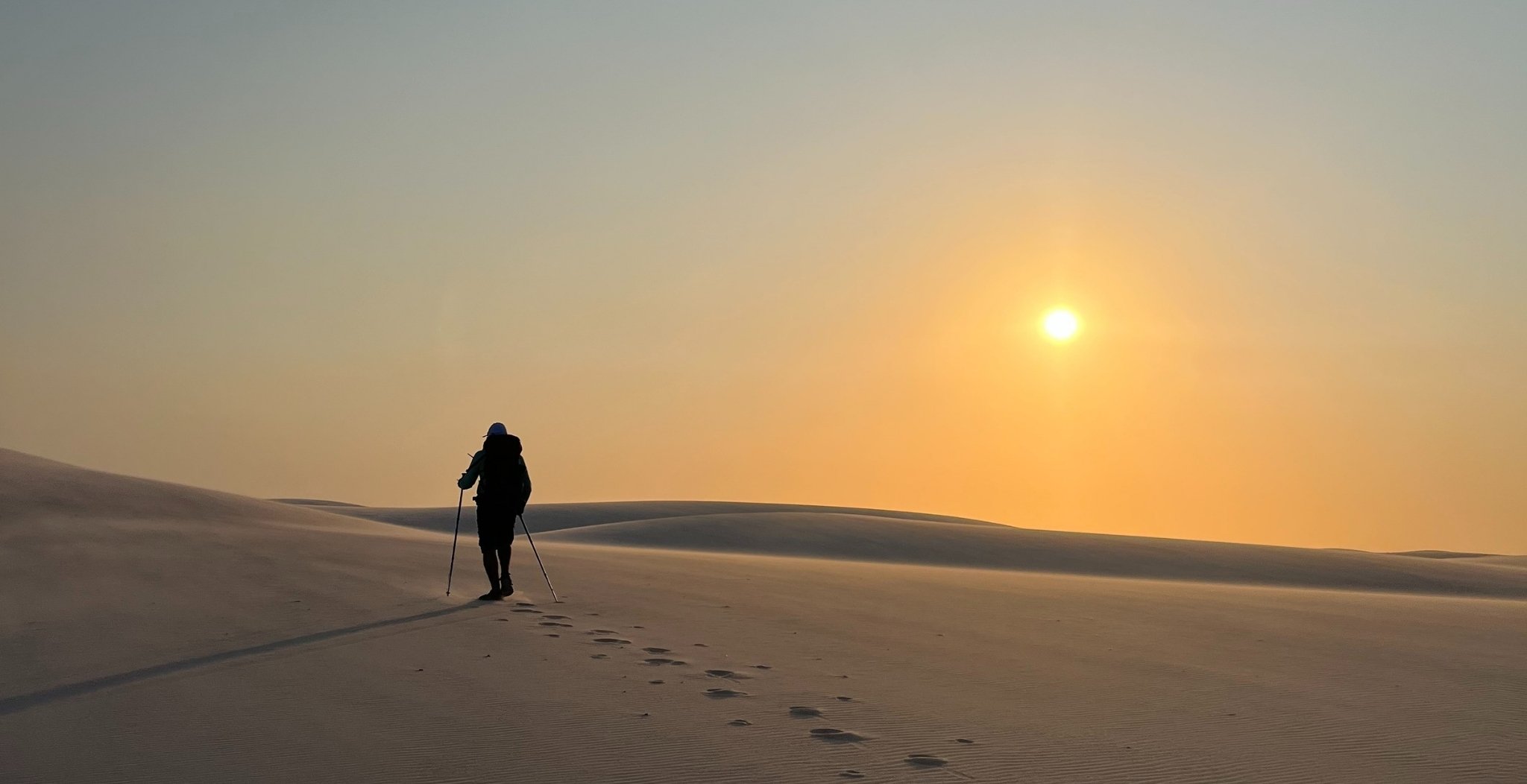 A lone hiker with trekking poles walks up a vast white sand dune toward a low golden sun on the lencois maranhenses trek, footprints trailing behind on the smooth sand