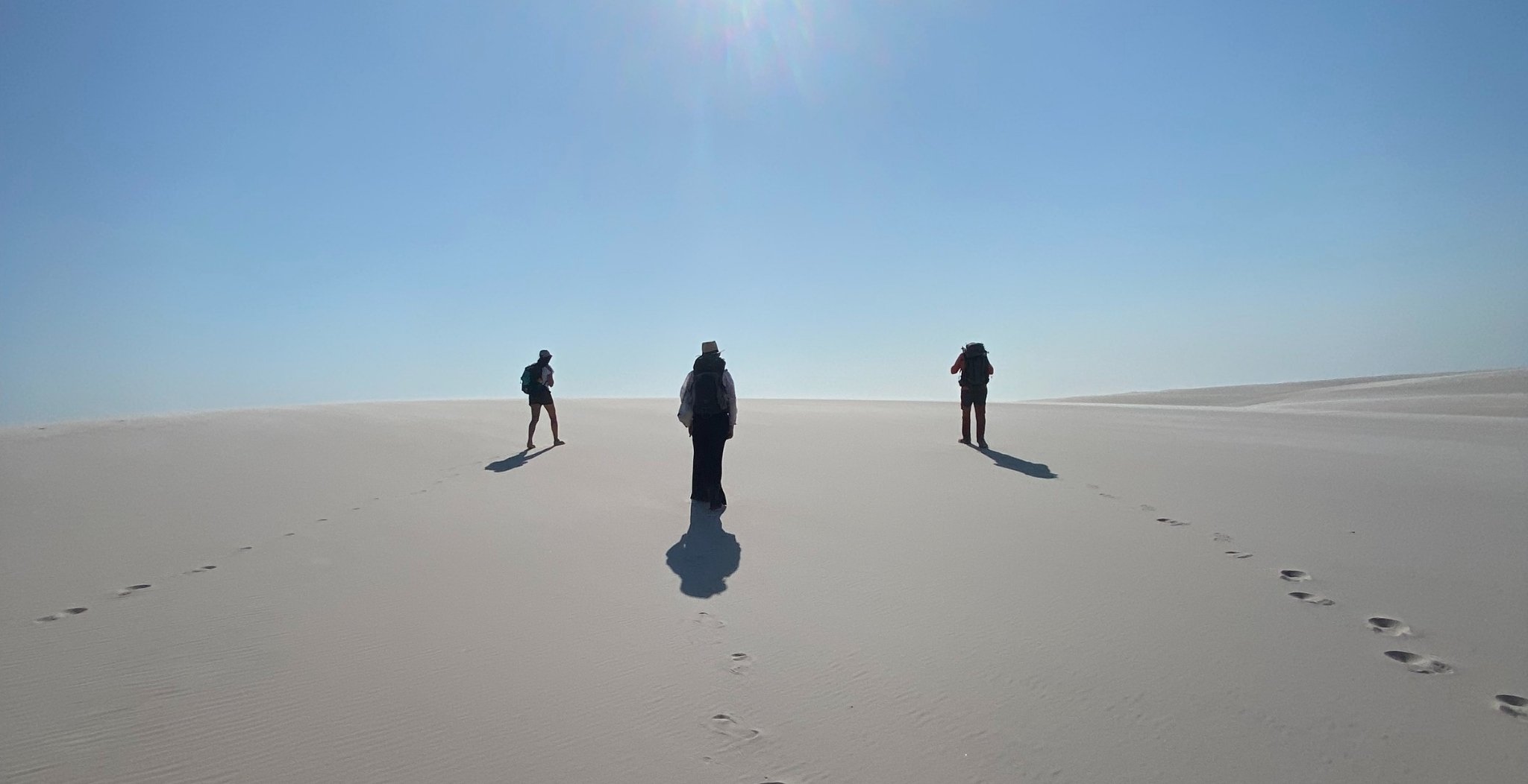 Three hikers with backpacks walk across a vast expanse of white sand dunes under a bright blue sky during a Lençóis Maranhenses trekking expedition, their footprints trailing behind them
