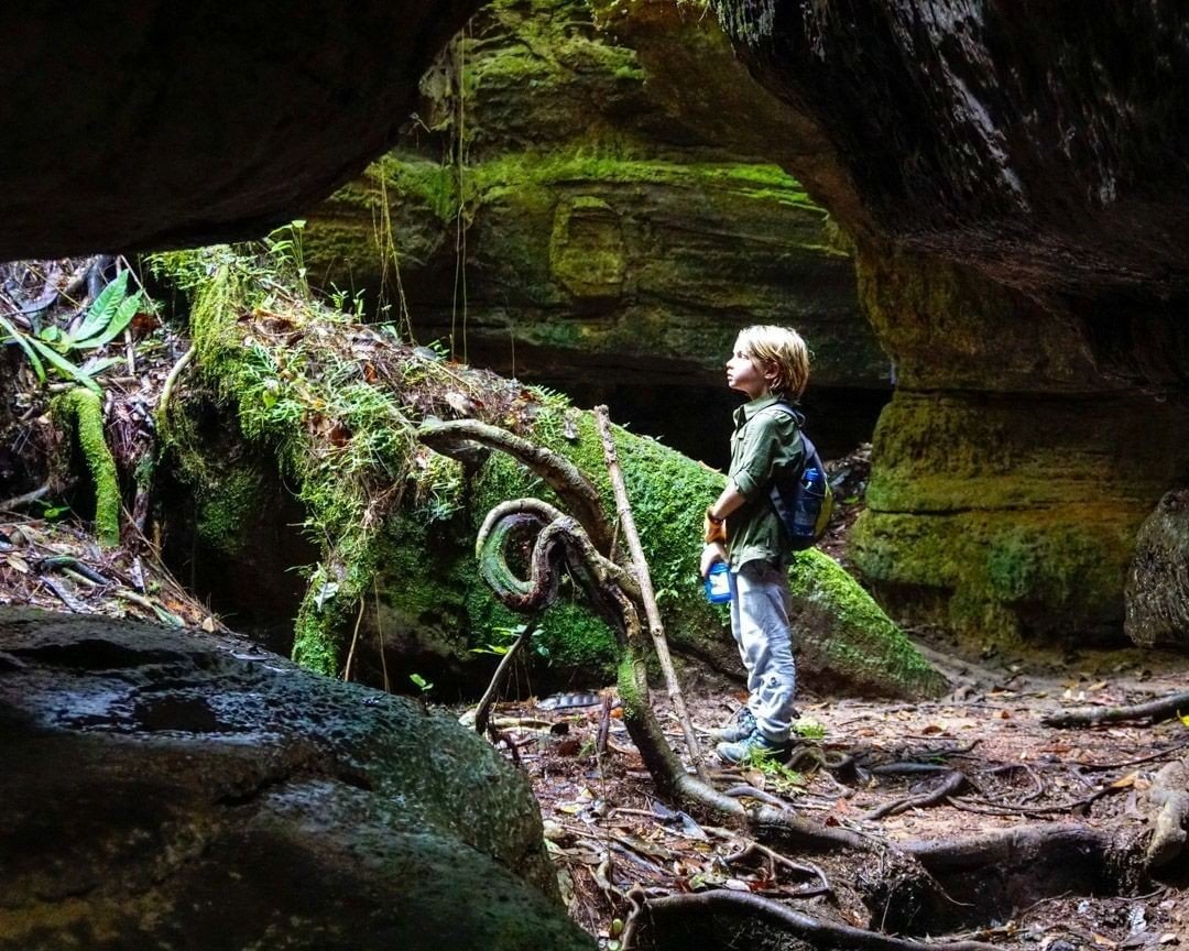 Een jong kind met een rugzakje kijkt vol ontzag omhoog in een met mos bedekte rotsachtige grot bij Mirante do Gavião, omringd door verweven wortels en weelderige begroeiing