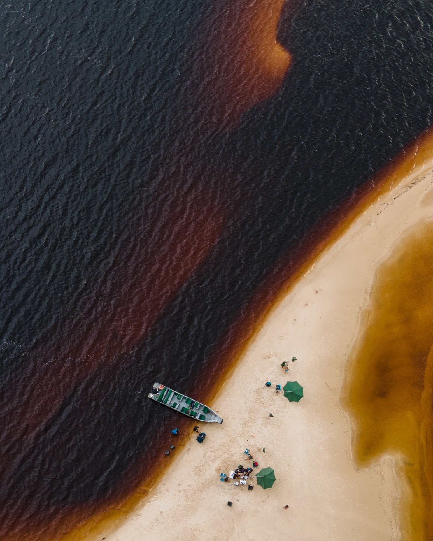 Twee rivieren, één zandbank, en een boot die duidelijk de juiste keuze maakte. Luchtfoto van een klein zandstrand aan de Rio Negro bij Mirante do Gavião, waar donker zwart water grenst aan een amberkleurige oever, met een houten boot aangemeerd aan de waterkant en twee groene parasols die bezoekers beschaduwen