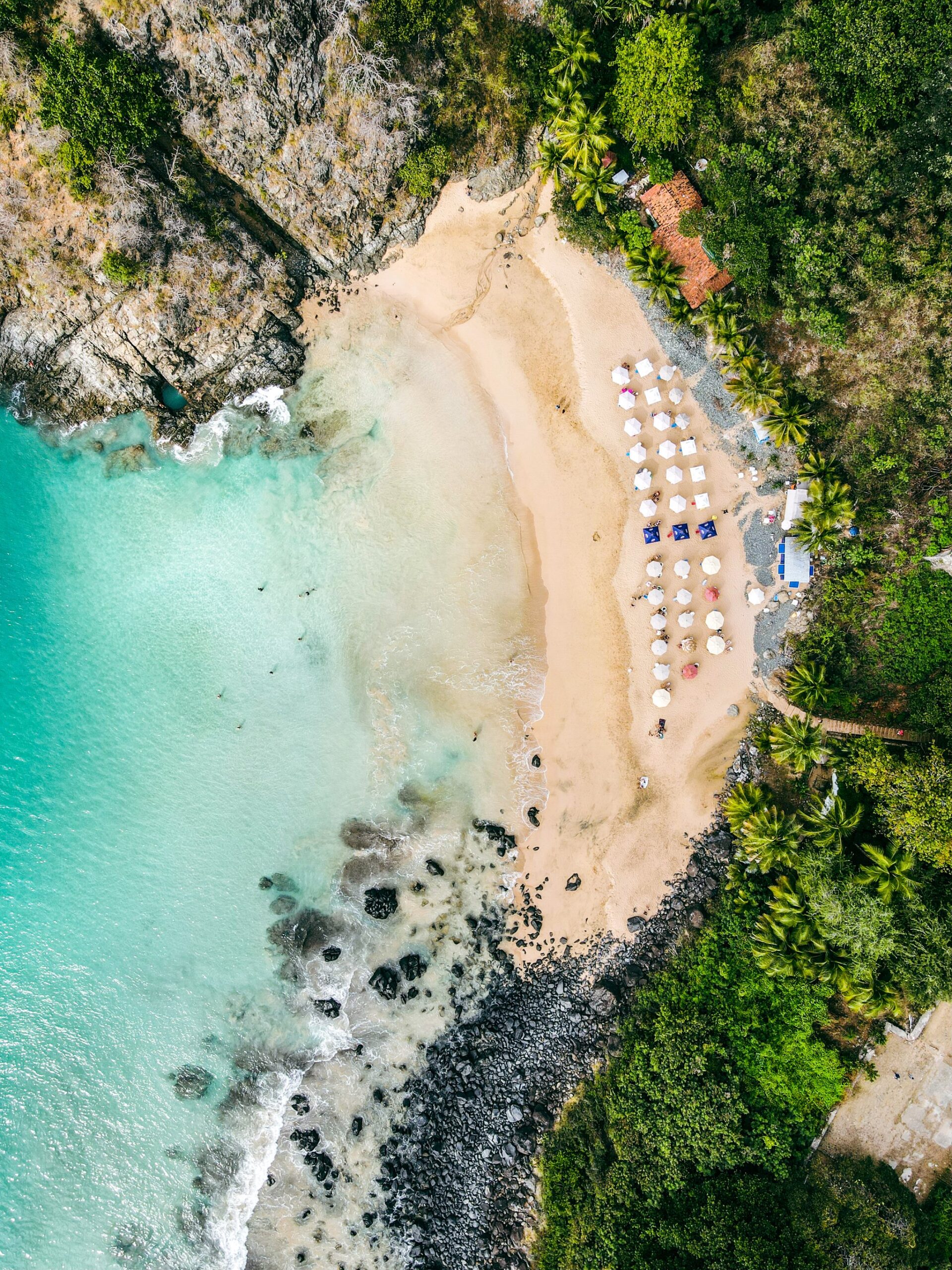 Your first beach dives happen somewhere that looks exactly like this. Aerial view of a small sandy cove in Fernando de Noronha, tucked between rocky cliffs and lush green vegetation, with rows of white beach umbrellas and turquoise water — the kind of sheltered bay where an Open Water diving course begins with calm beach training dives