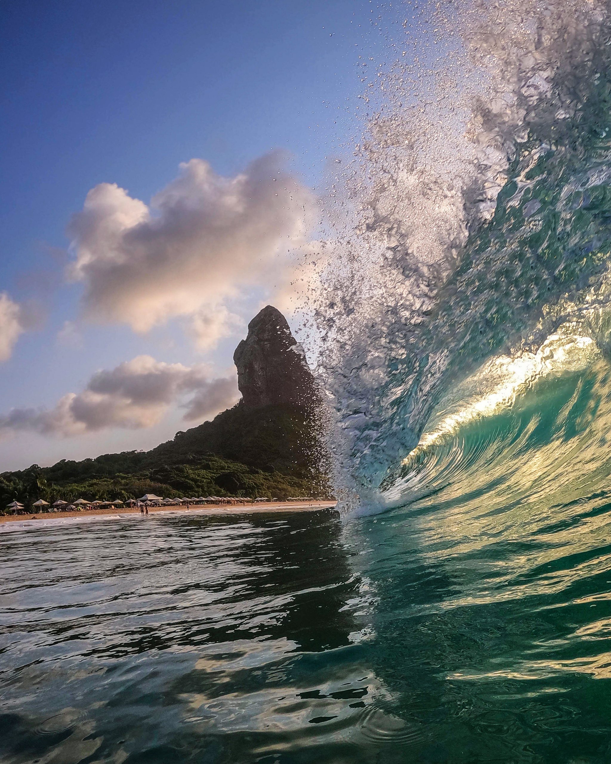 The ocean here has opinions, and it shares them loudly. A turquoise barrel wave curls and splashes in the foreground of an Open Water diving course in Fernando de Noronha, with the iconic Morro do Pico rock pinnacle and a palm-fringed beach visible behind it under a partly cloudy blue sky.