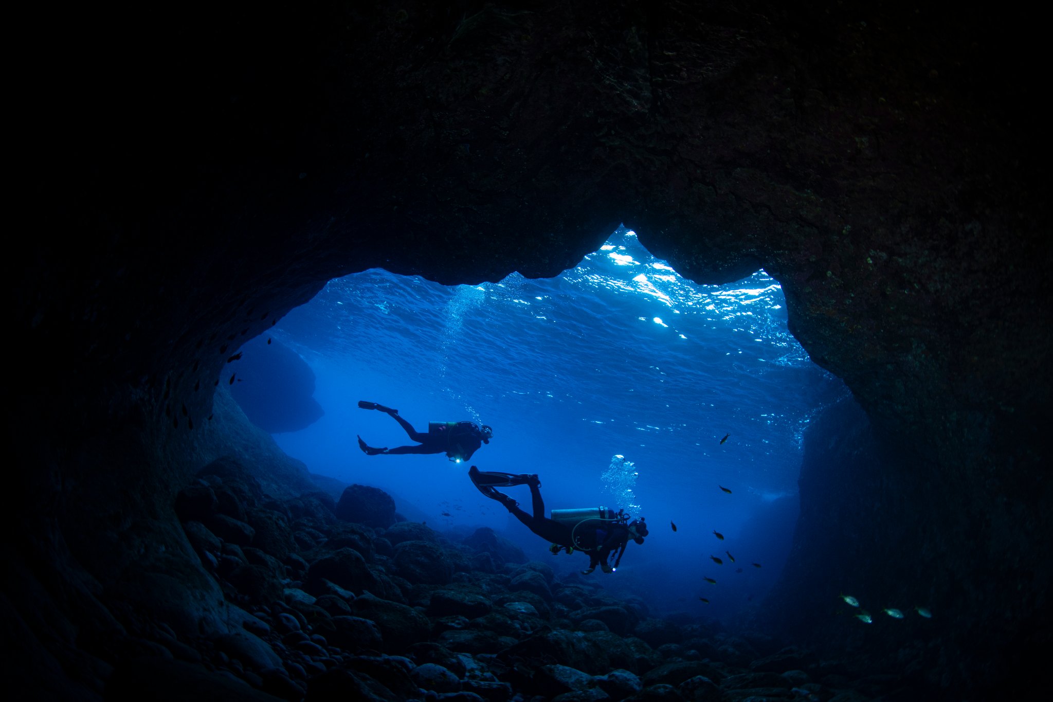 Two scuba divers silhouetted against deep blue water inside a rocky underwater cave during an Open Water diving course, with sunlight filtering through the cave mouth and small fish visible nearby
