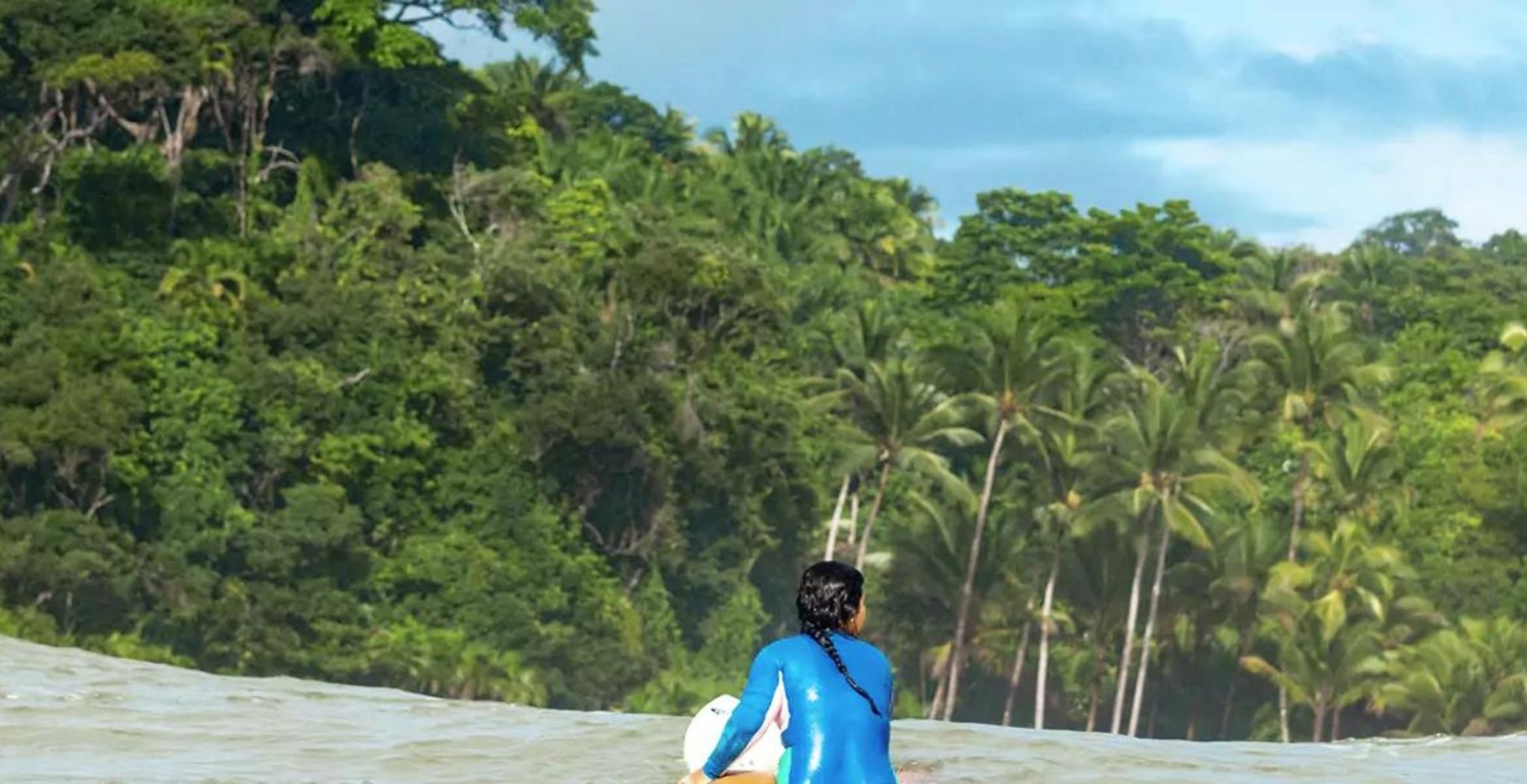 A surfer in a blue wetsuit carrying a surfboard wades into the ocean during surf lessons in Itacaré, with dense Atlantic Forest and palm trees rising behind the shoreline